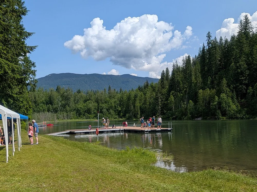People gathering on a wooden dock and shoreline by a lake, surrounded by green trees and mountains under a partly cloudy sky.