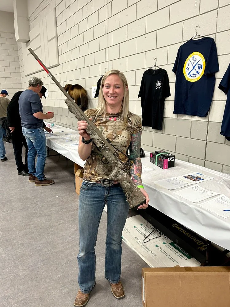 A woman smiling and holding a large rifle at an indoor event. People are seen in the background, and various shirts are hanging on the wall.