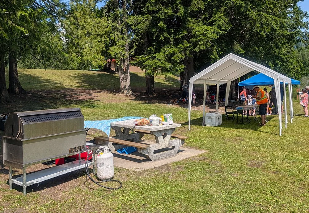 Park setting with a barbecue grill, a picnic table, and a white canopy tent hosting people.