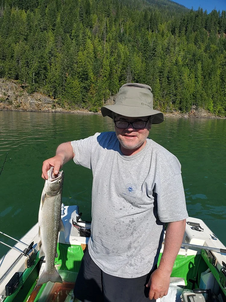 A man wearing a wide-brimmed hat, glasses, and a gray t-shirt holding a fish on a boat in a lake with a forested mountain backdrop.