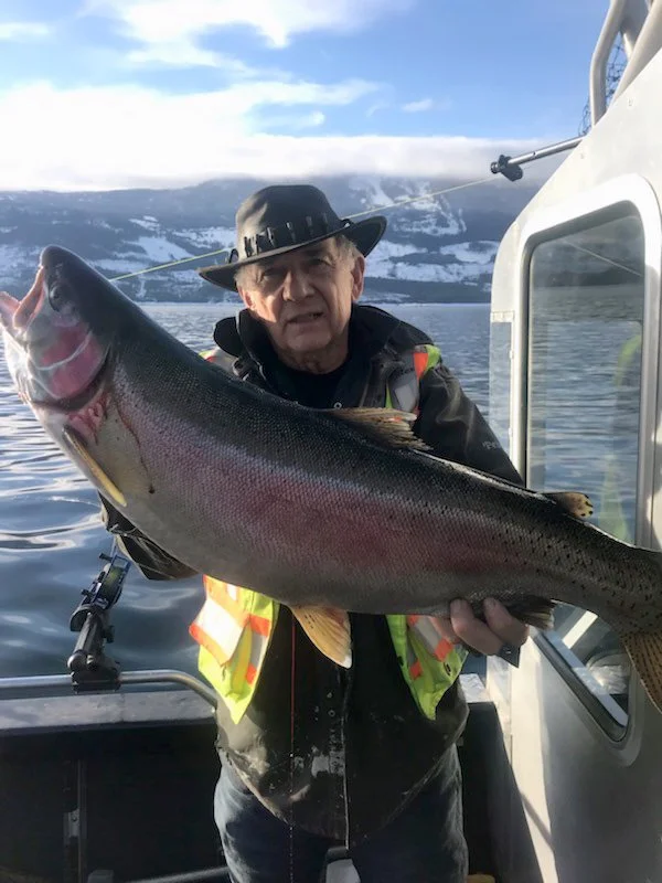 An elderly man in a black hat and jacket holding a large fish on a boat with a scenic mountain and lake background.