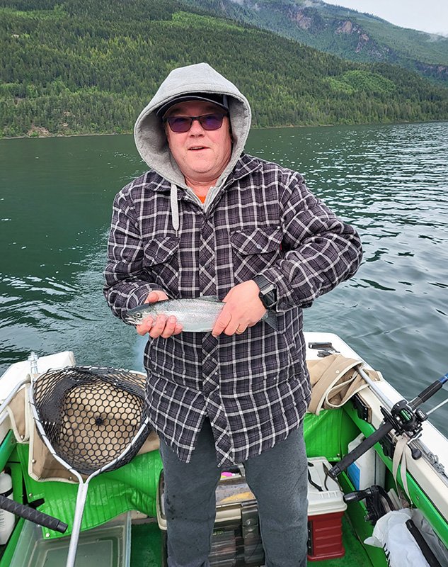 A woman in a checkered jacket, gray hoodie, and sunglasses is on a boat holding a fish, with a lake, green forest, and hills in the background.