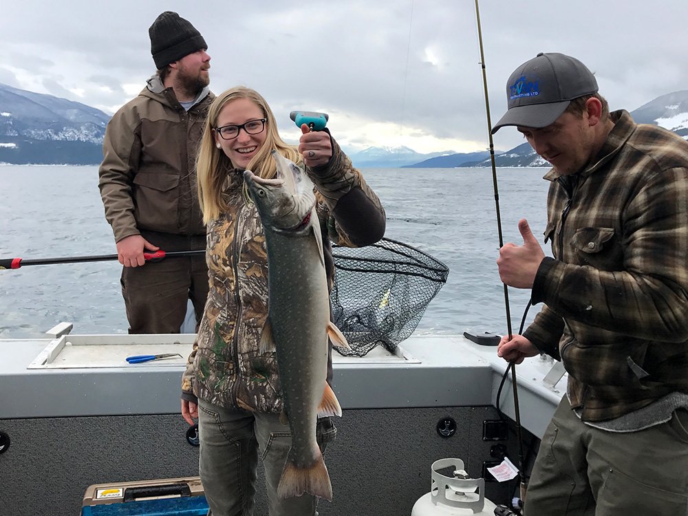 Three people on a boat in a lake, with mountains in the background, celebrating a successful fishing trip. One woman holds a large fish and smiles, while two men are nearby, one giving a thumbs up and the other holding a fishing rod.