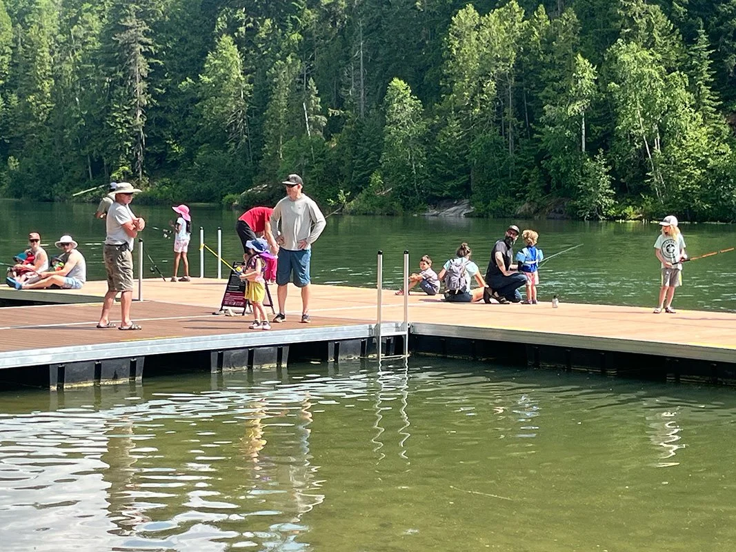 People fishing and sitting on a wooden dock by a lake surrounded by trees.