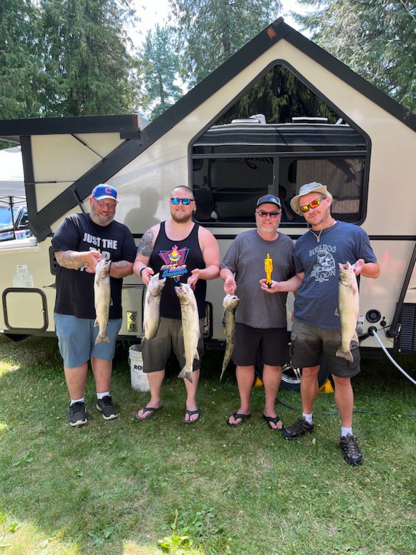Four men standing in front of an RV, each holding a large fish they caught, with two of them holding a fishing tool. They are outdoors in a wooded area on grass, smiling and wearing casual summer clothes.