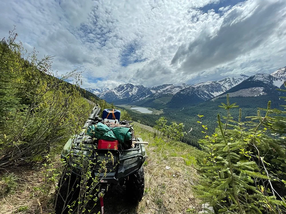 An all-terrain vehicle parked on a grassy hillside with backpacks and gear, overlooking a mountain valley with snow-capped peaks, a lake, and a cloudy sky.