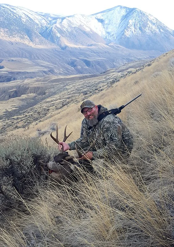 A man in camouflage clothing kneels in tall grass, holding the antlers of a deer he has hunted, with mountains and snow in the background.