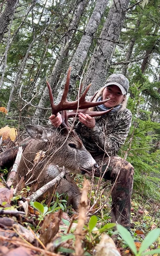 Woman in camouflage clothing with a cap, smiling and holding antlers of a large deer lying on the forest ground.