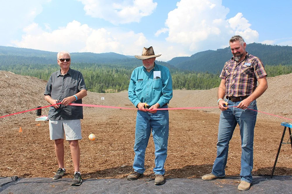 Three men standing outdoors in front of a large dirt area, holding a pink ribbon for a ribbon-cutting ceremony, with mountains and blue sky with scattered clouds in the background.