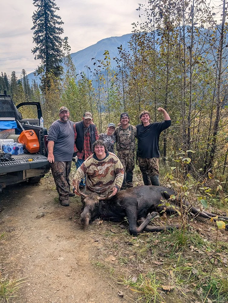 Group of six hunters posing with a deceased black bear in a forested area during daytime, with a mountain in the background and hunting gear on a nearby truck.