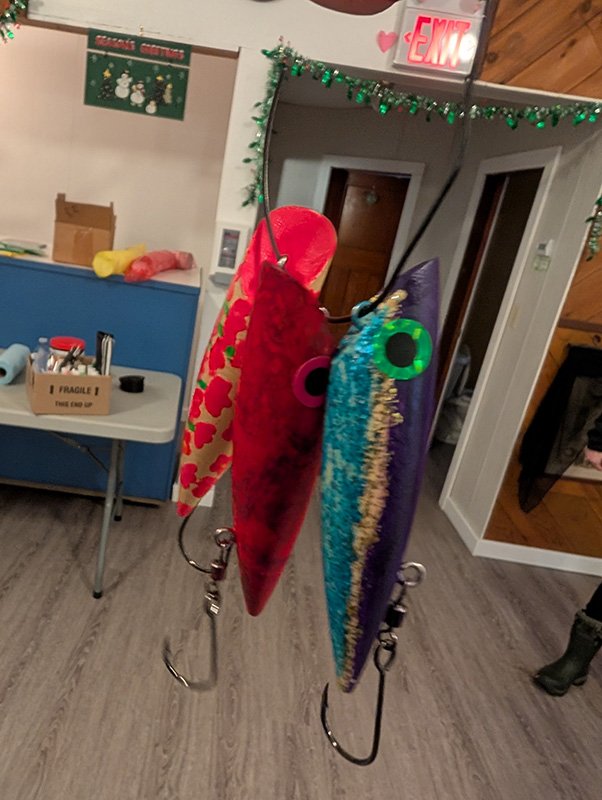 Two colorful fishing lures hanging in an indoor setting with a table, a cardboard box, and decorated walls in the background.