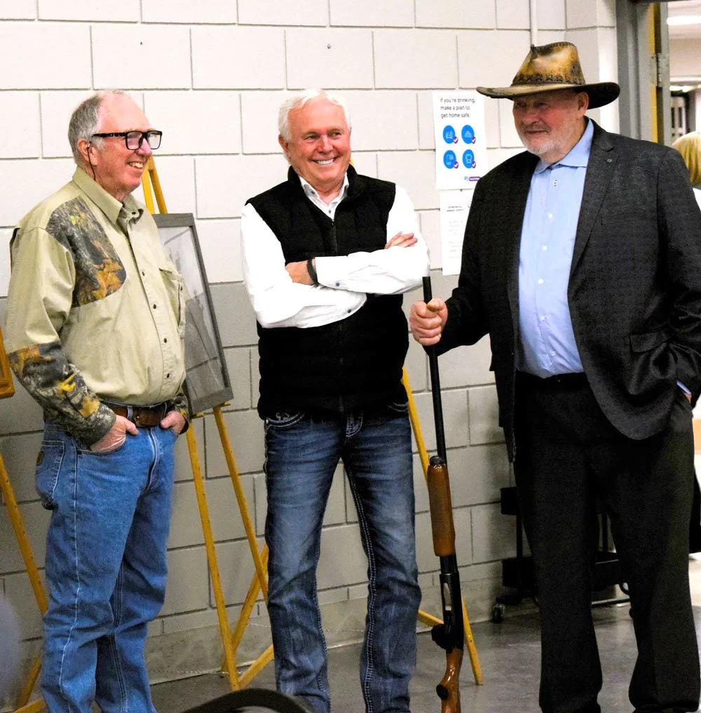 Three men standing indoors, smiling, one holding a rifle, another wearing a cowboy hat, people in the background, and posters on the wall.