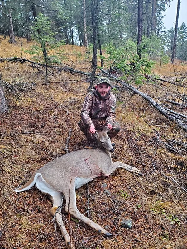 A man in camouflage clothing squats in a forested area next to a recently hunted deer lying on the ground. The deer appears to be a young female with a small set of antlers. The forest has tall trees, ferns, and fallen branches.