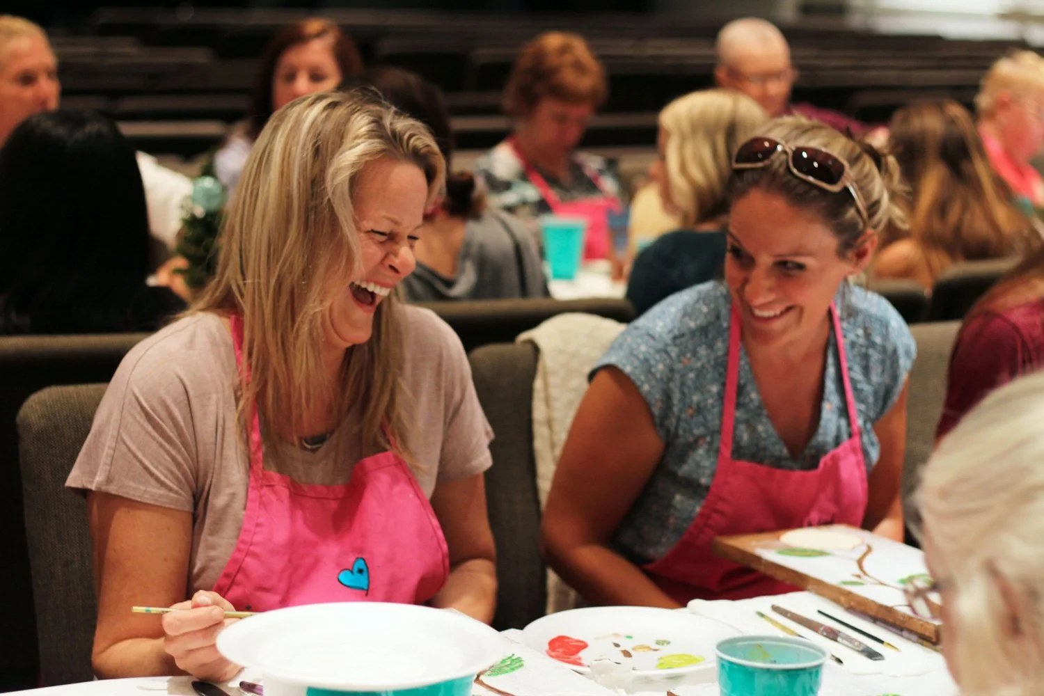 Two women laughing and painting at a table during a group activity. They are wearing pink aprons and surrounded by painting supplies in an indoor setting with other participants in the background.