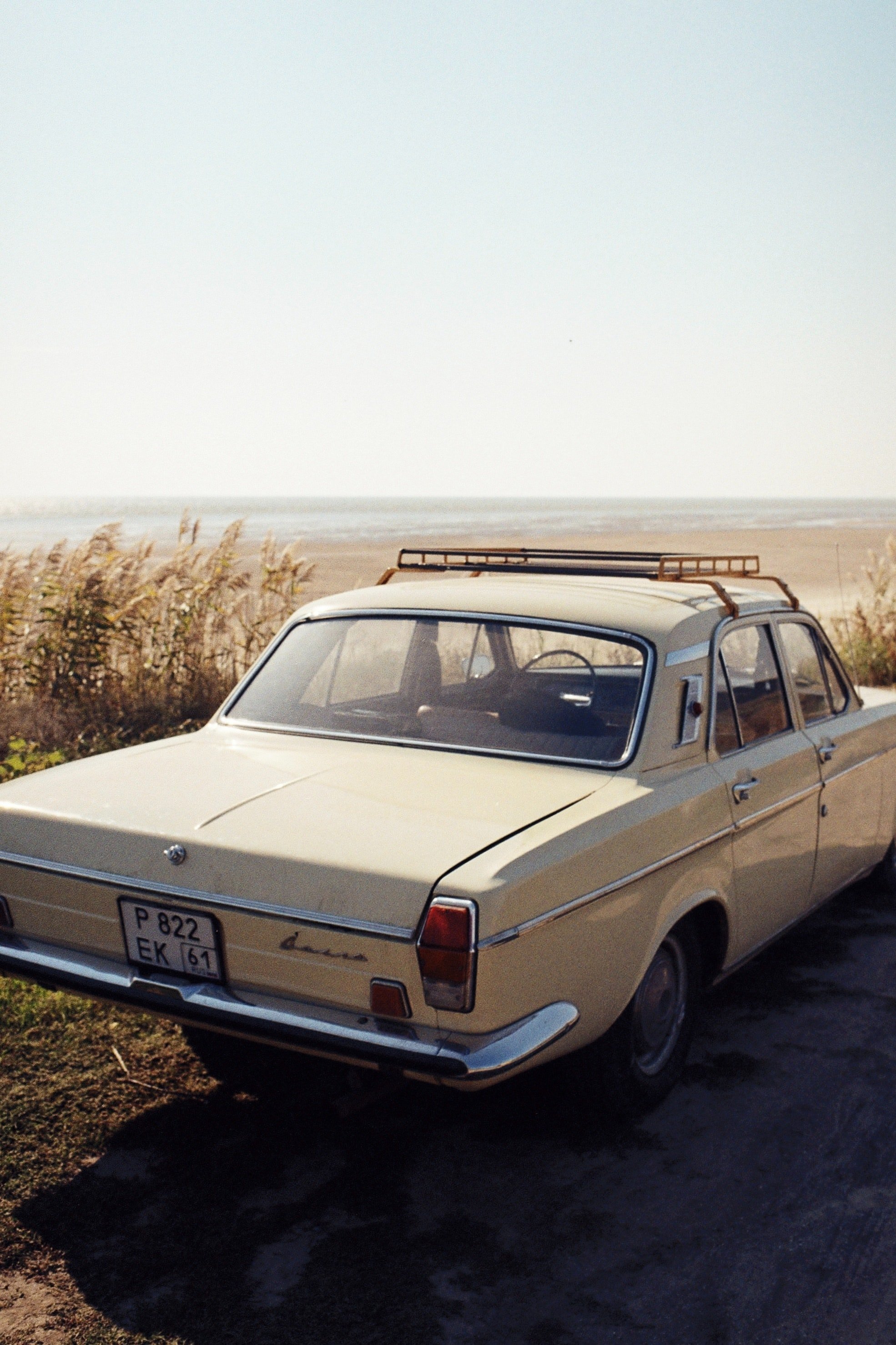 A vintage beige car parked outdoors near a beach with a grassy area and a clear sky in the background.