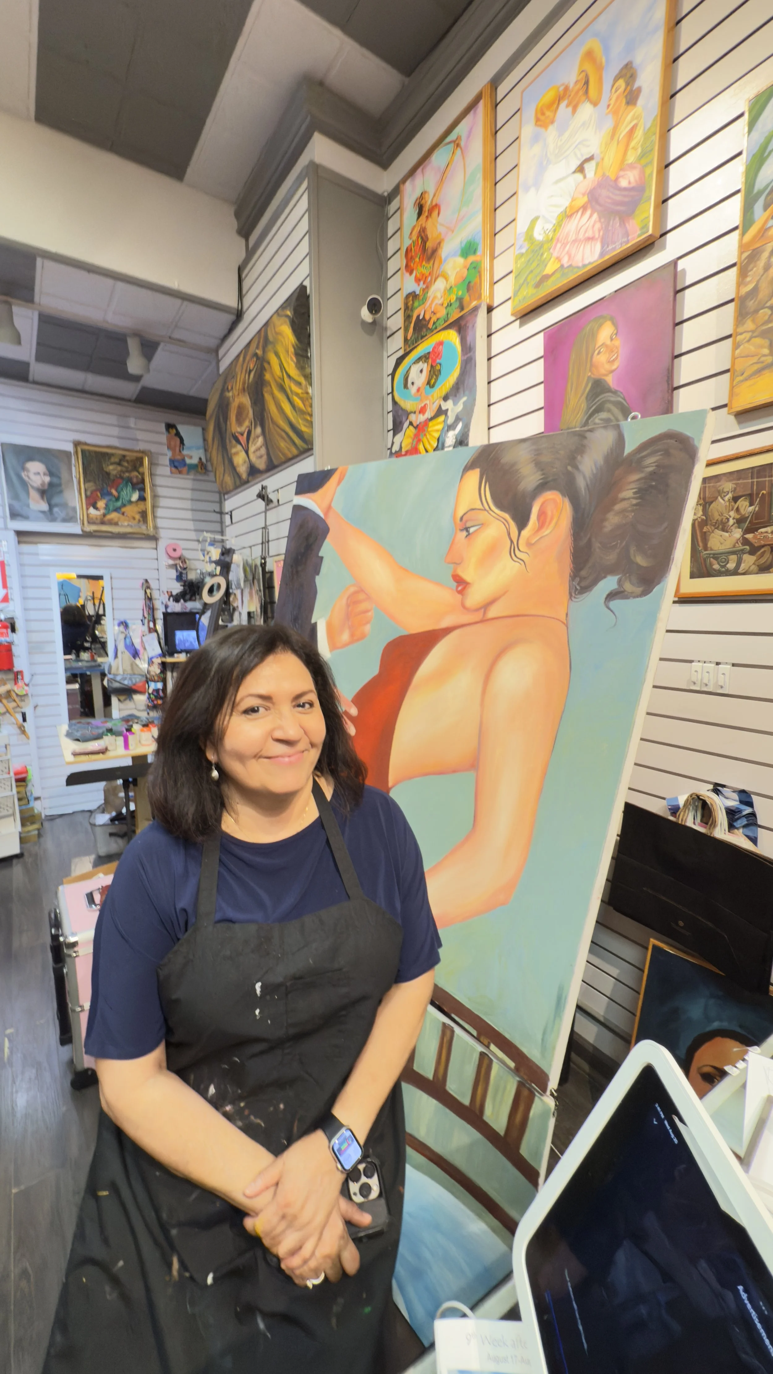A woman standing inside an art gallery, smiling, wearing a black apron with paint splatters, and a smartwatch, beside a large painting of a woman flexing her arm.