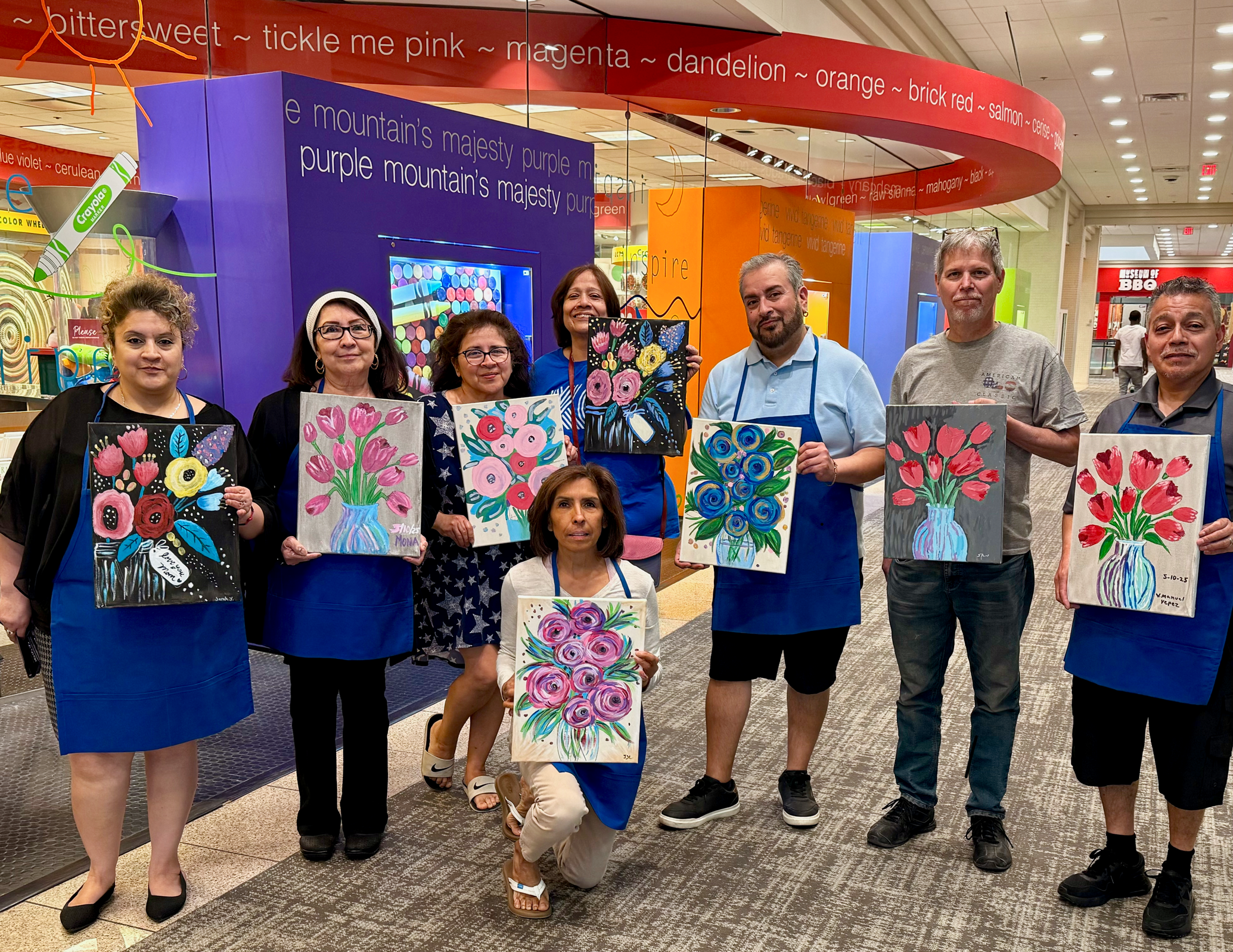 Group of people in a shopping mall holding colorful paintings of flowers and vases, standing in front of a vibrant, display-themed wall.