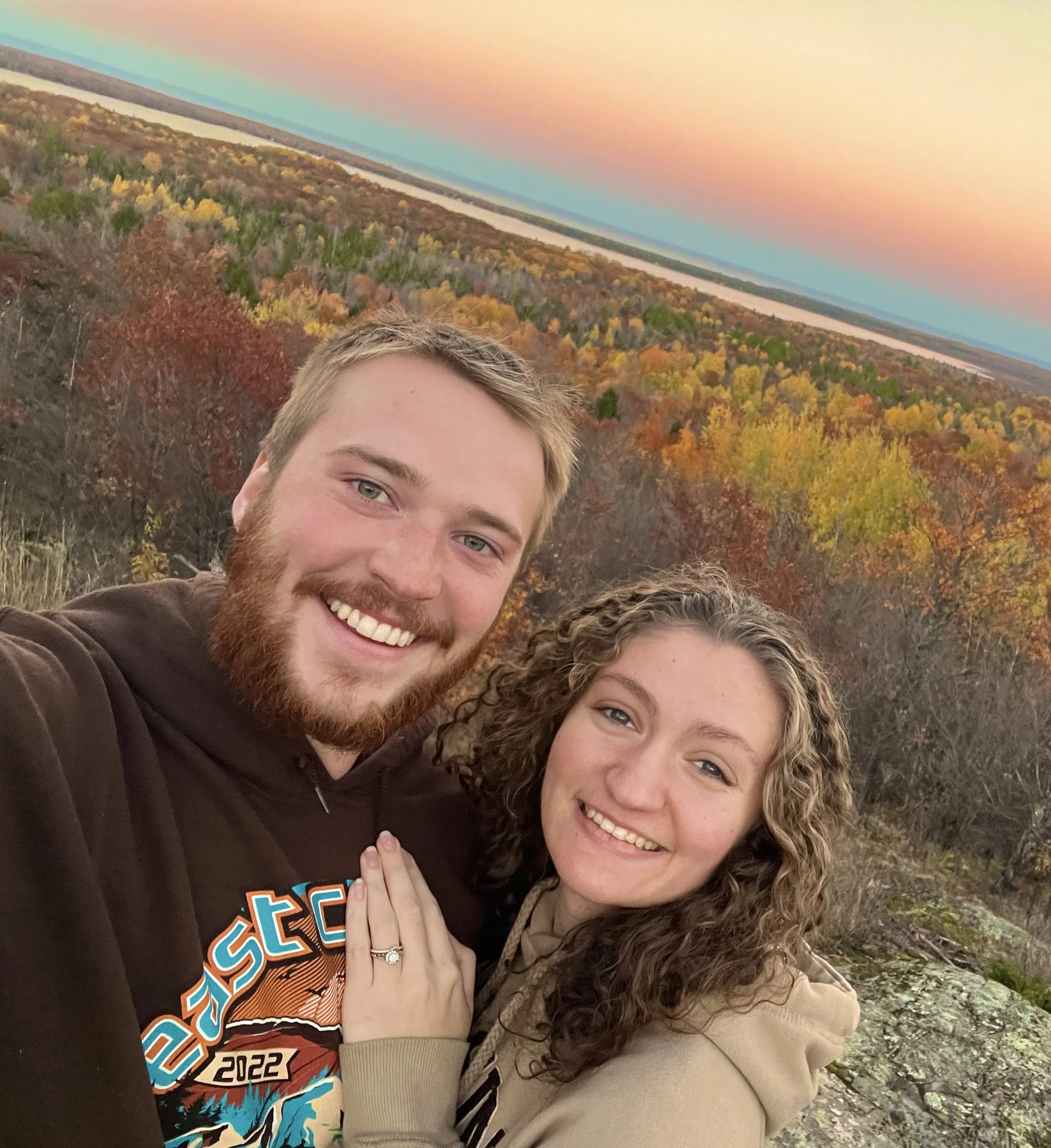 A happy couple taking a selfie outdoors during autumn with a colorful forest and river in the background.