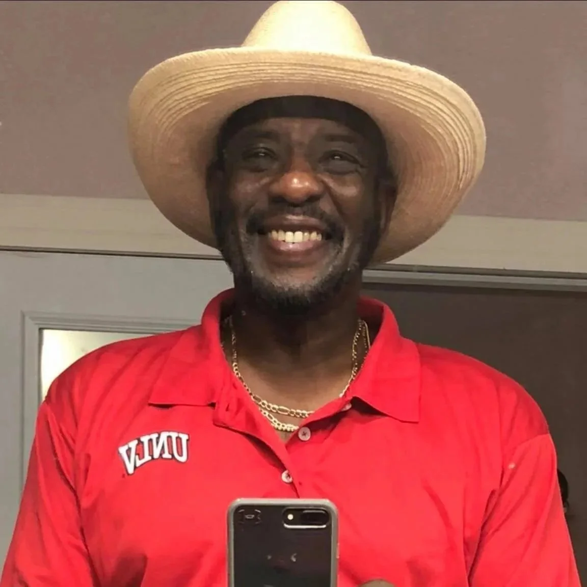 A smiling man wearing a large straw hat, a red UNEA polo shirt, a gold chain, and taking a selfie indoors.