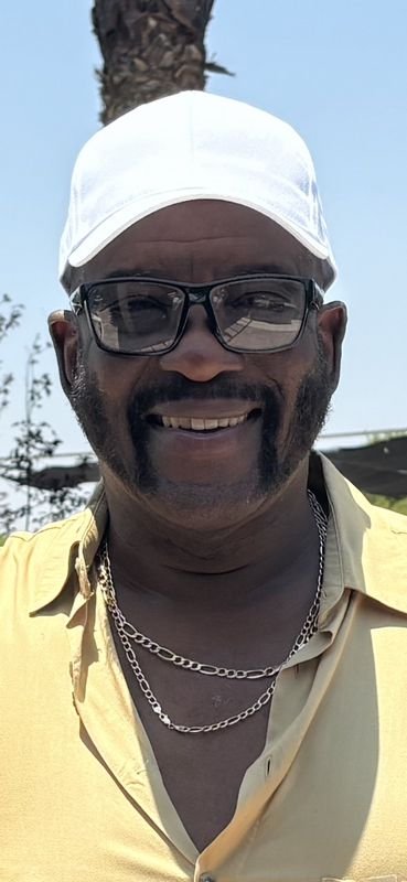 A man smiling outdoors, wearing sunglasses, a white cap, a yellow shirt, and layered gold chains around his neck, with a palm tree and blue sky in the background.