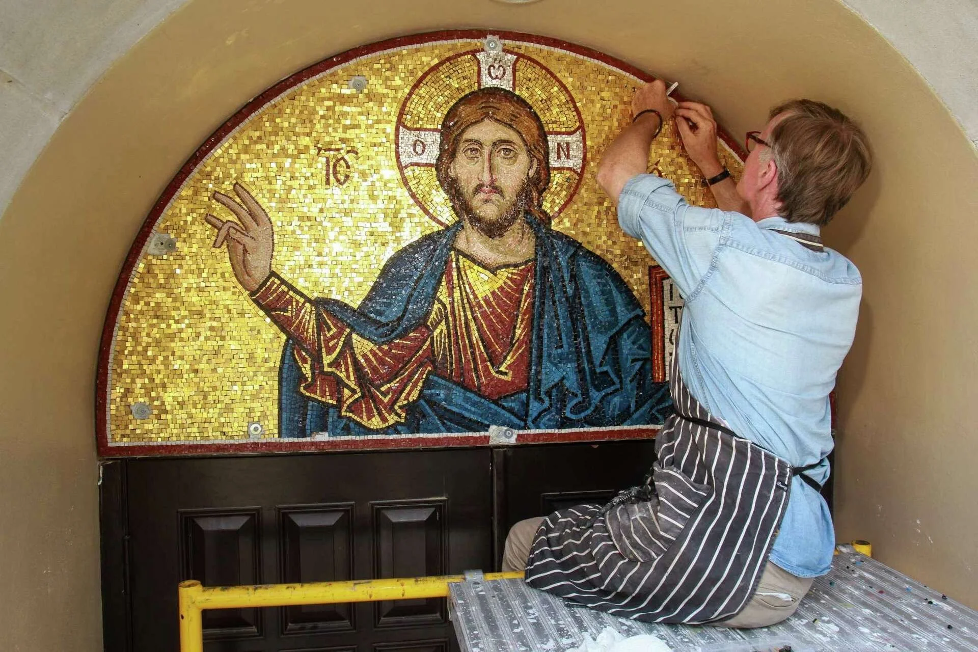 A person working on a mosaic of Jesus Christ, sitting on a platform while placing mosaic tiles on the artwork.