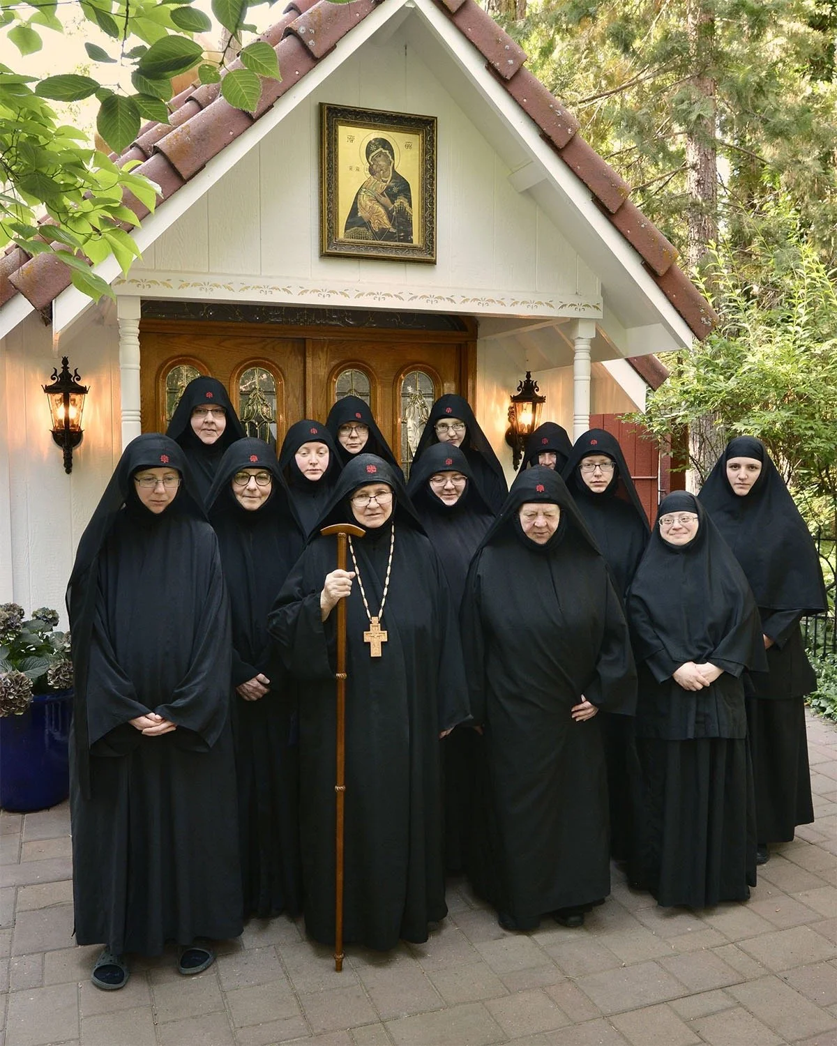 A group of nuns dressed in black habits standing in front of a small church or chapel with a religious icon above the door, during daytime.