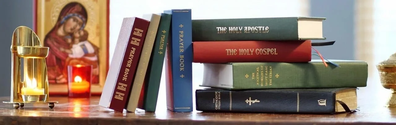 Stack of religious books on a wooden table, with icons and candles in the background.