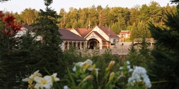 Large house with arched entrance surrounded by trees and shrubs, with mountains in the background and flowers in the foreground.