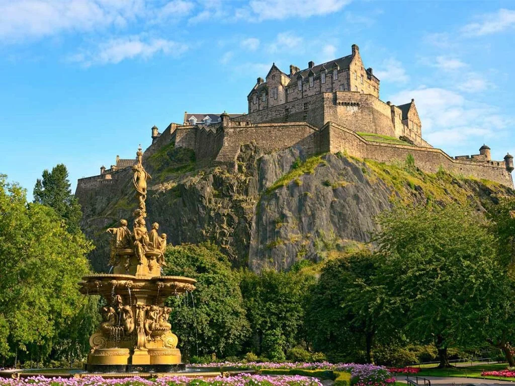 A castle on a rocky hilltop with a fountain and lush green trees in the foreground under a partly cloudy blue sky.