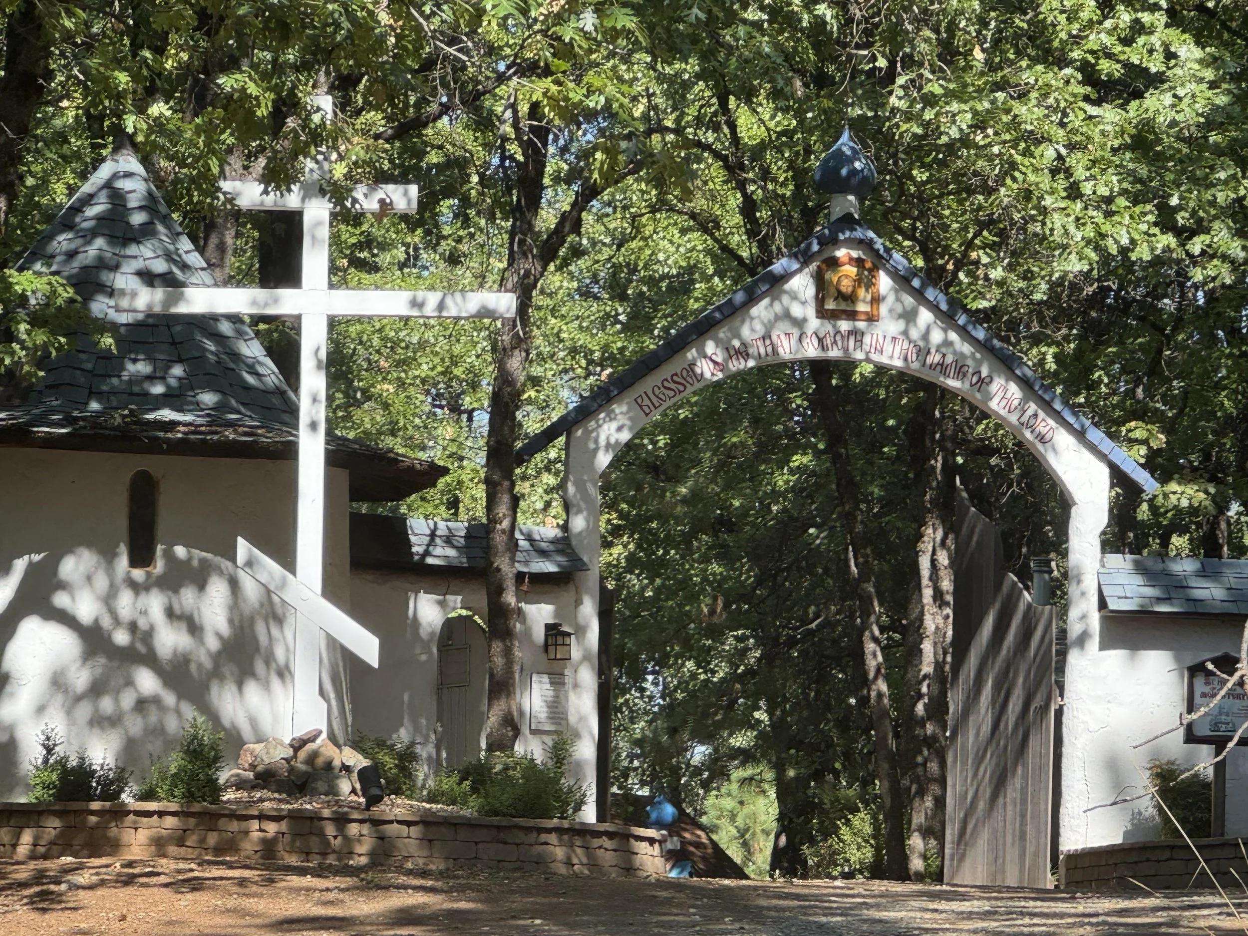 View of a small white chapel with a cross and a miniature house, surrounded by trees, with an arched sign that reads 'Blessed are those that come in the name of the Lord'.