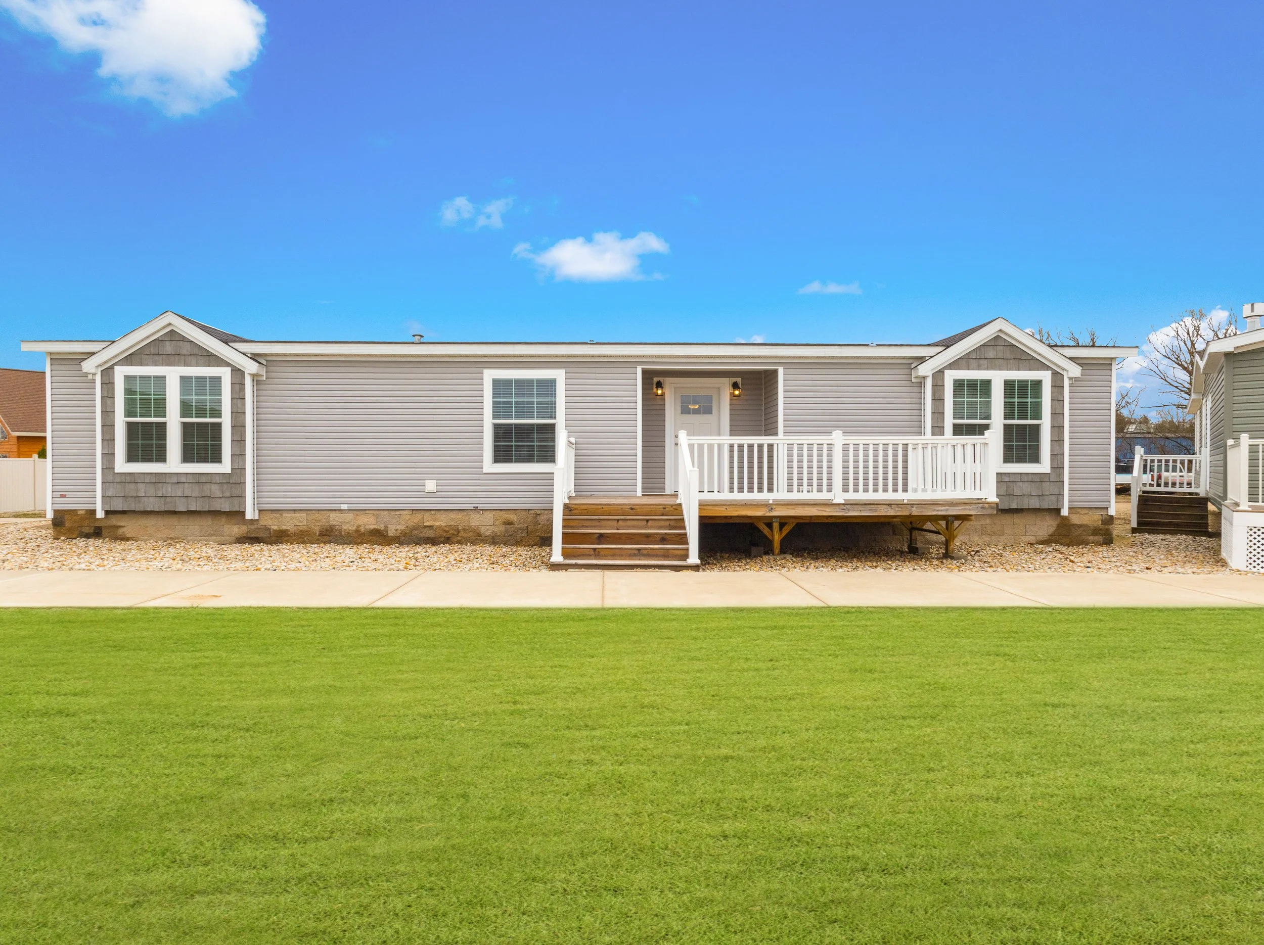 A light gray, modern mobile home with a small porch and white railing, two windows with white frames, and two small gable roofs on each side, set against a bright blue sky with some clouds and surrounded by a grassy lawn and gravel pathway.