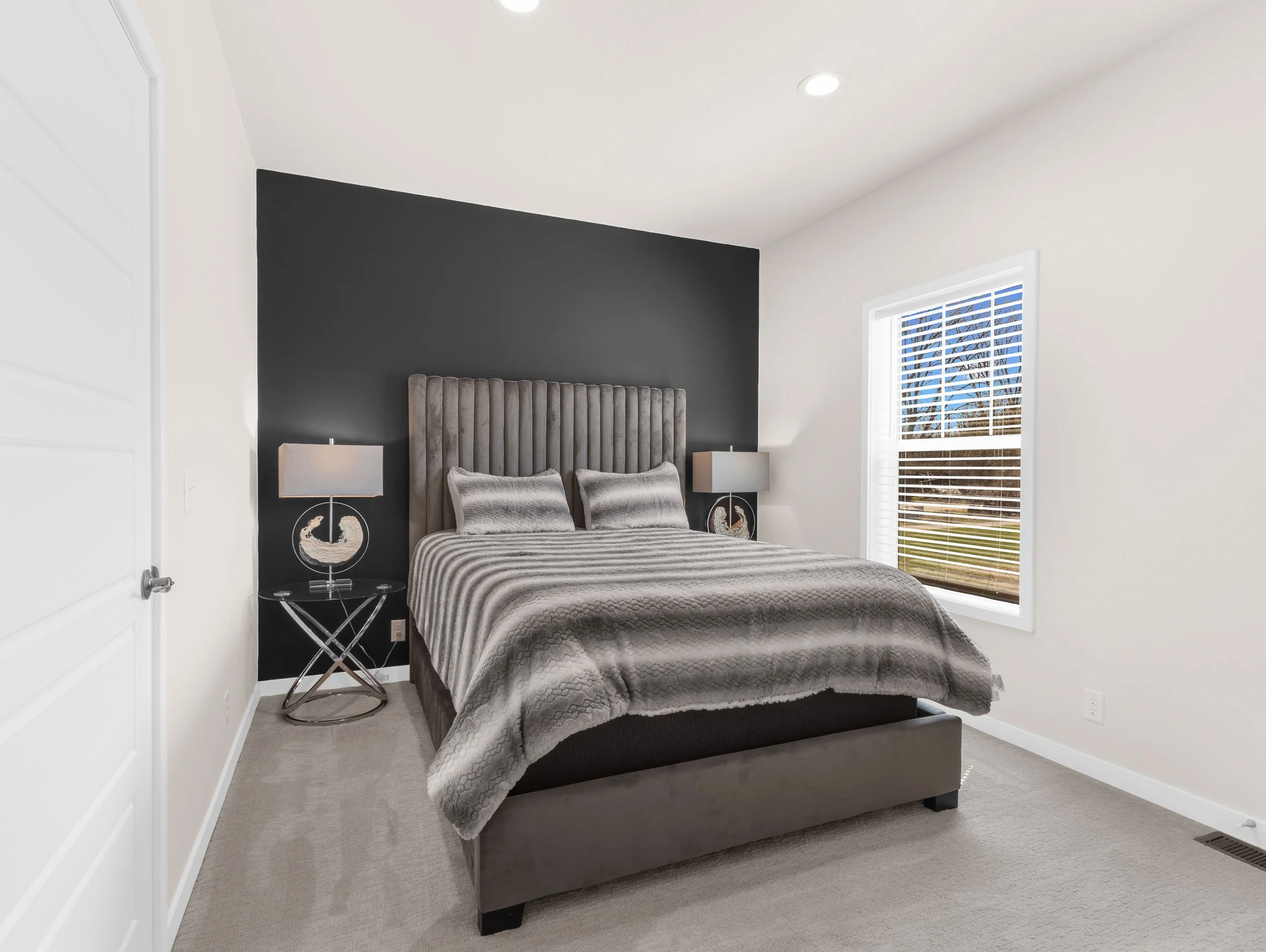 A bedroom featuring a bed with a high, tufted, gray upholstered headboard, two matching gray pillows, and a quilted gray bedspread. On either side of the bed are nightstands with modern lamps. The room has a dark accent wall behind the bed, white wal