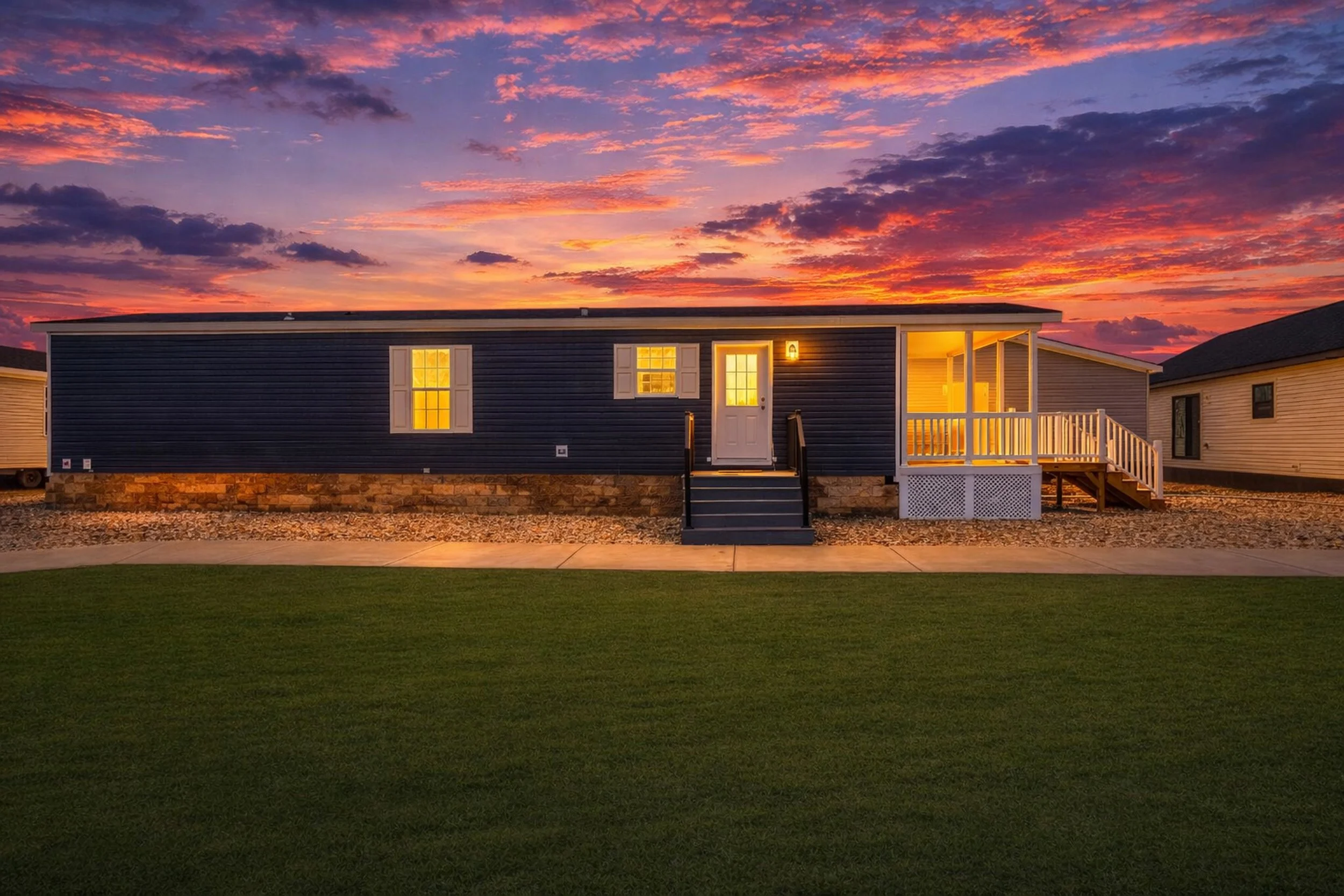 A small house with dark blue siding, white window shutters, and a porch with white railing, illuminated by warm yellow lights, set against a vibrant sunset sky with pink, purple, and orange clouds, and a grassy area in the foreground.