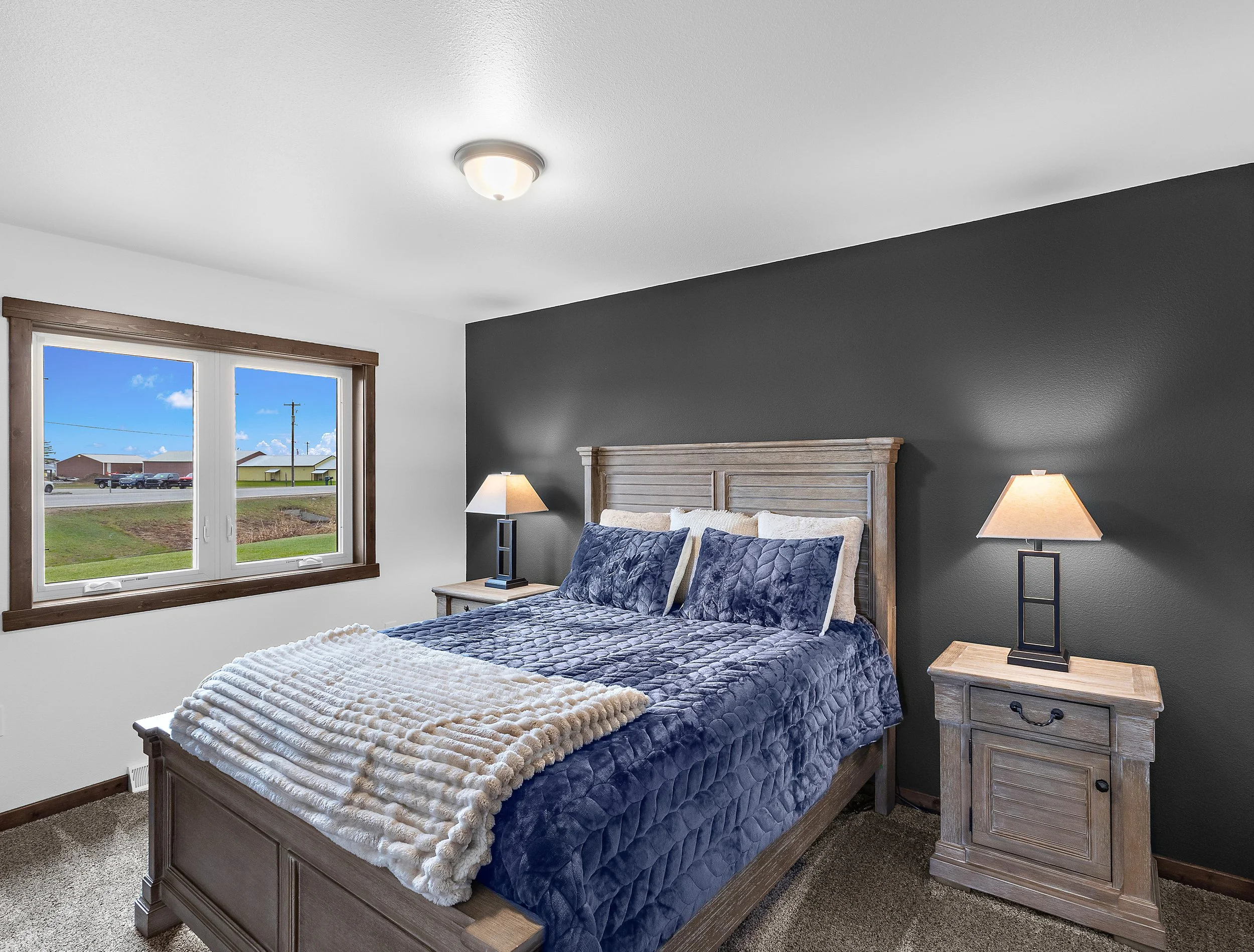 Bedroom with wooden bed frame, blue quilt, white blanket, two lamps on nightstands next to dark gray accent wall, and window showing a cloudy sky and parked cars outside.
