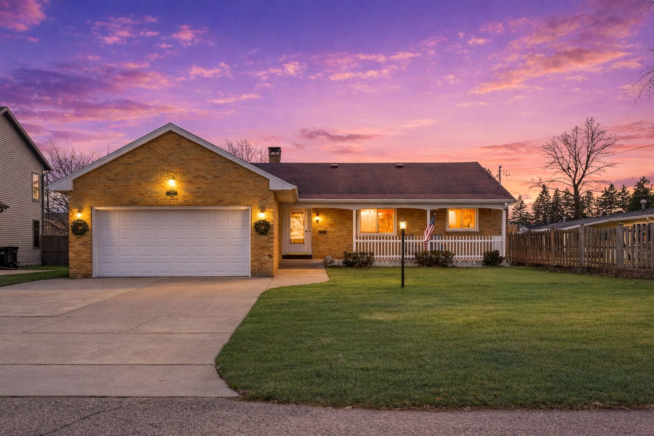 A brick house with an attached garage illuminated by outdoor lights during sunset, with a front yard, a sidewalk, a mailbox, and neighboring houses visible, under a colorful sky with purple and pink clouds.