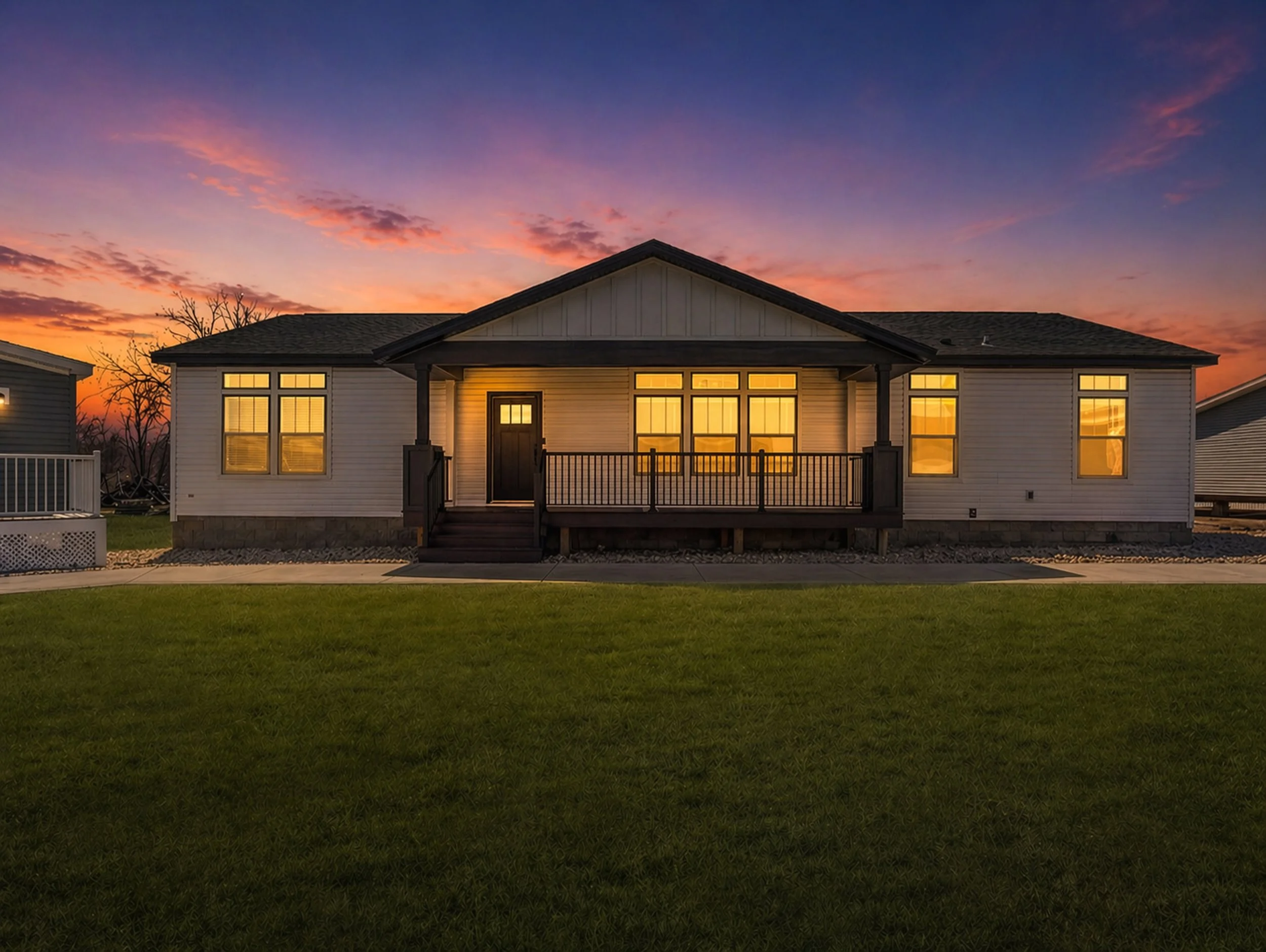 A house at sunset with a front porch, multiple large windows, and interior lights on. The sky is colorful with pink, purple, and orange hues, and there is a grassy lawn in the foreground.
