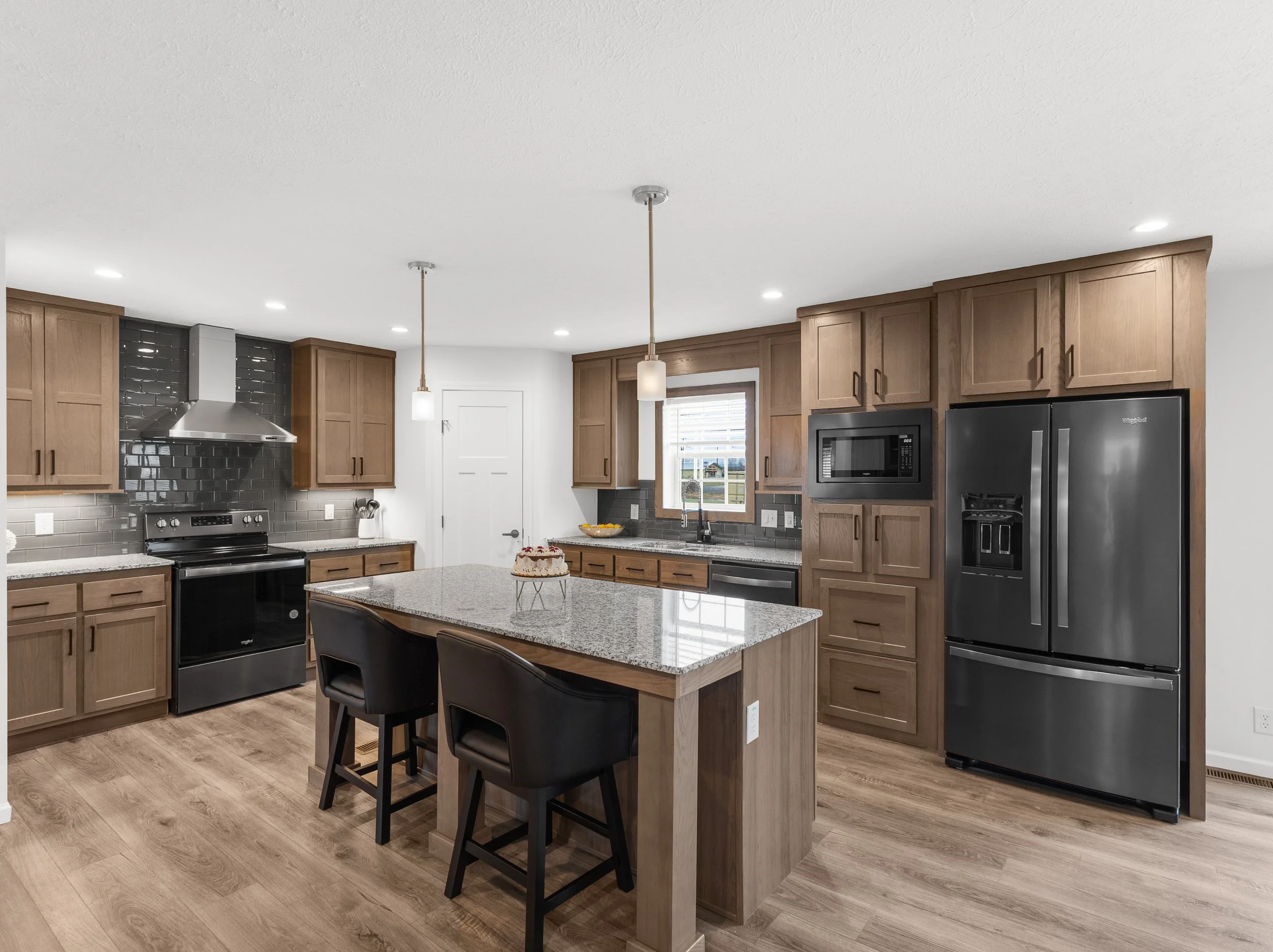 Modern kitchen with wooden cabinets, stainless steel appliances, a granite island, black chairs, pendant lights, and a window with blinds.