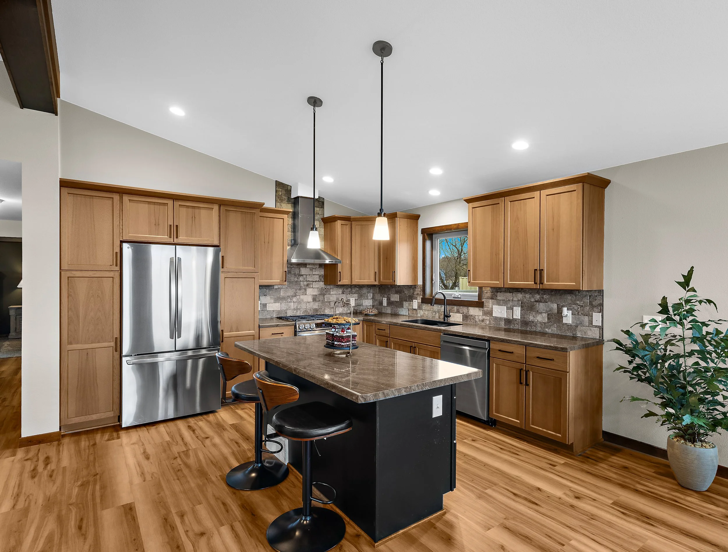 Modern kitchen with wooden cabinets, stainless steel refrigerator, brick backsplash, island with bar chairs, hanging pendant lights, and a window with a view of trees outside.