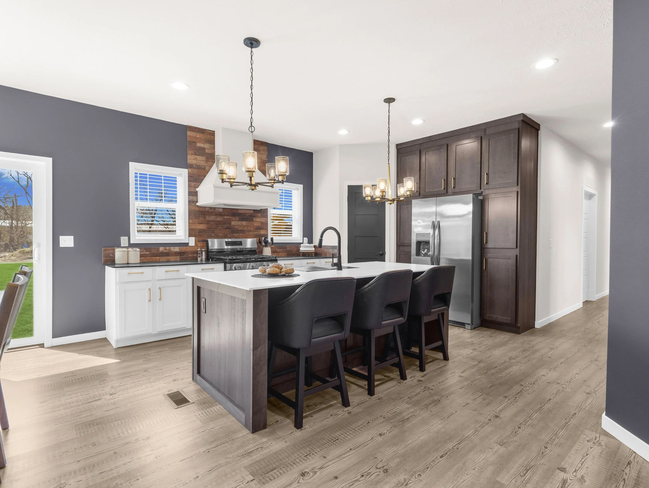 Modern kitchen with white island, dark wood cabinets, stainless steel refrigerator, brick accent wall, pendant and recessed lighting, large window with blinds, light wood flooring.