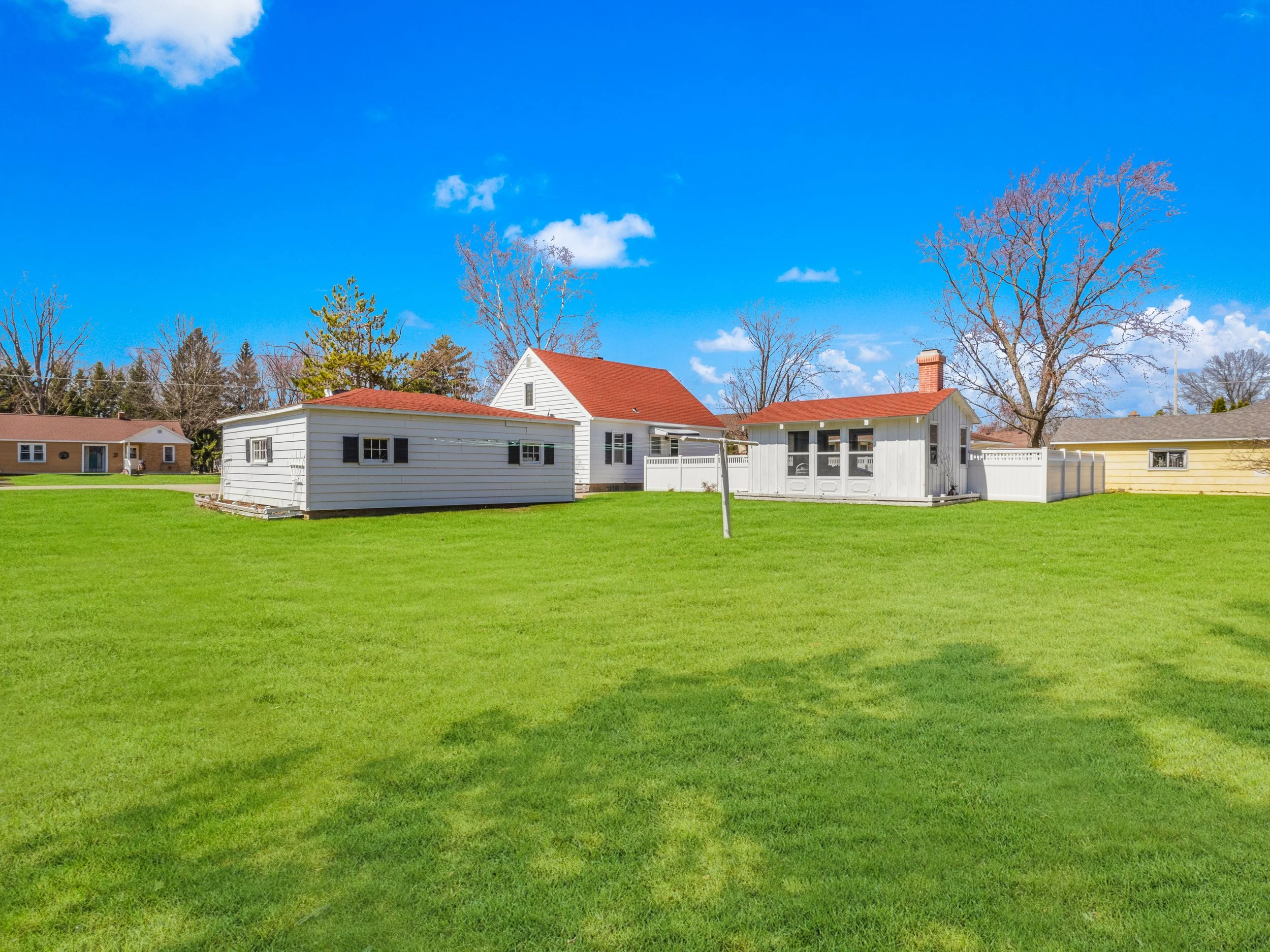 A backyard with bright green grass, several houses with white and yellow siding and red roofs, and leafless trees under a blue sky with a few clouds.