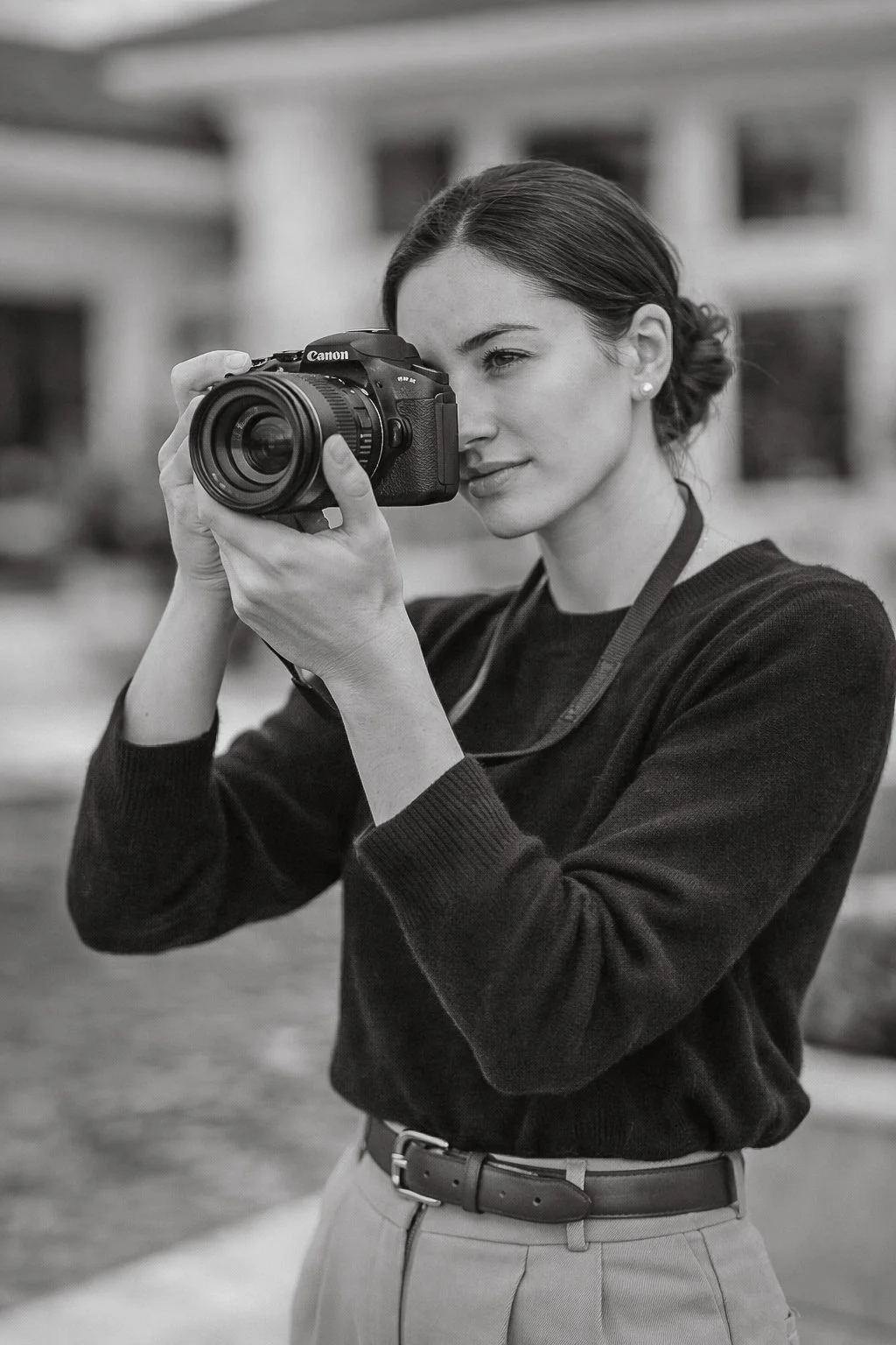 Black and white photo of a woman with dark hair holding a camera up to her eye, outdoors.