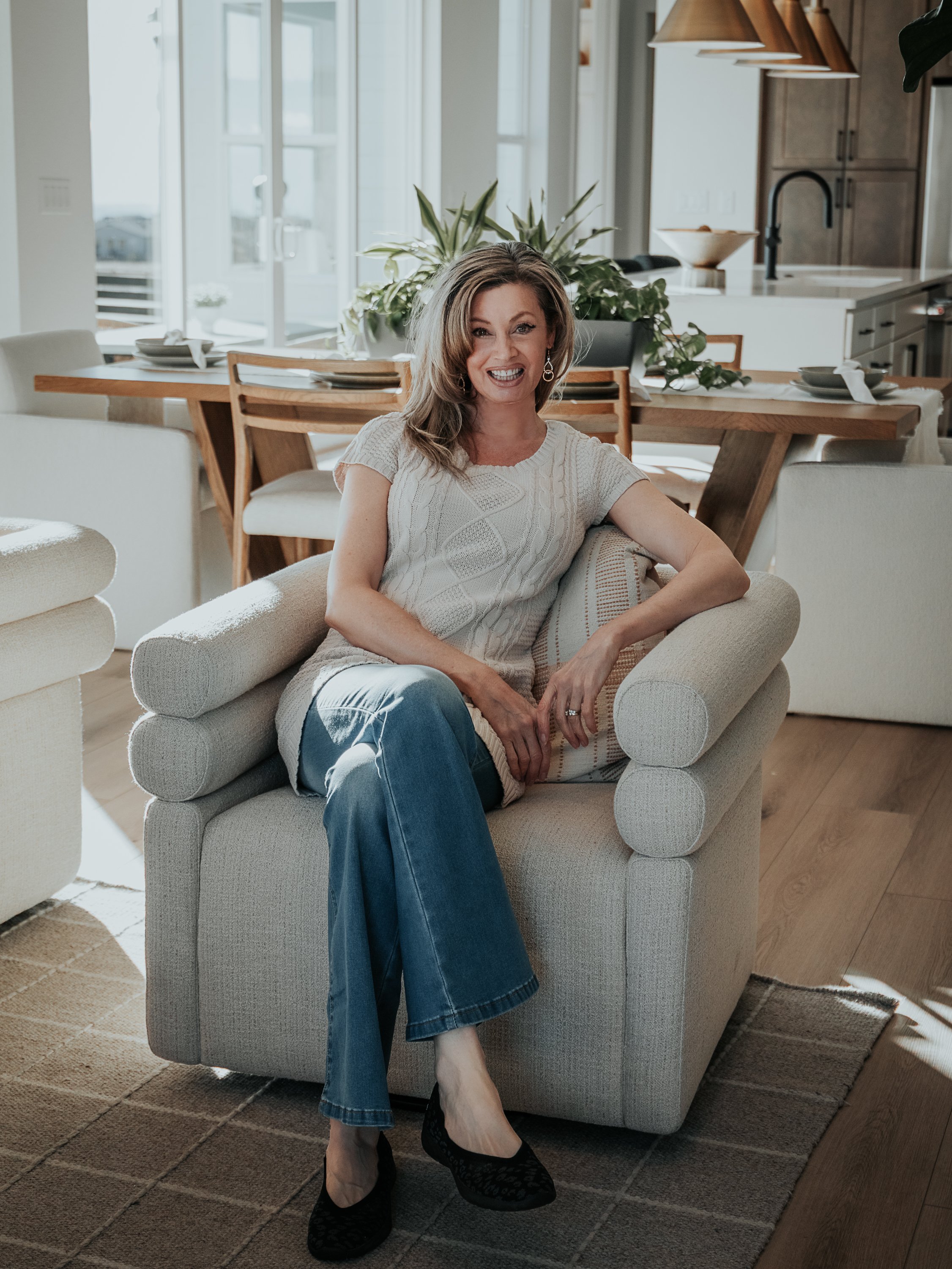 A woman sitting on a beige armchair in a modern, well-lit dining area. She is smiling, wearing a light-colored knit top, blue jeans, and black high heels. Behind her is a wooden dining table set with plates and silverware, with large windows and gree