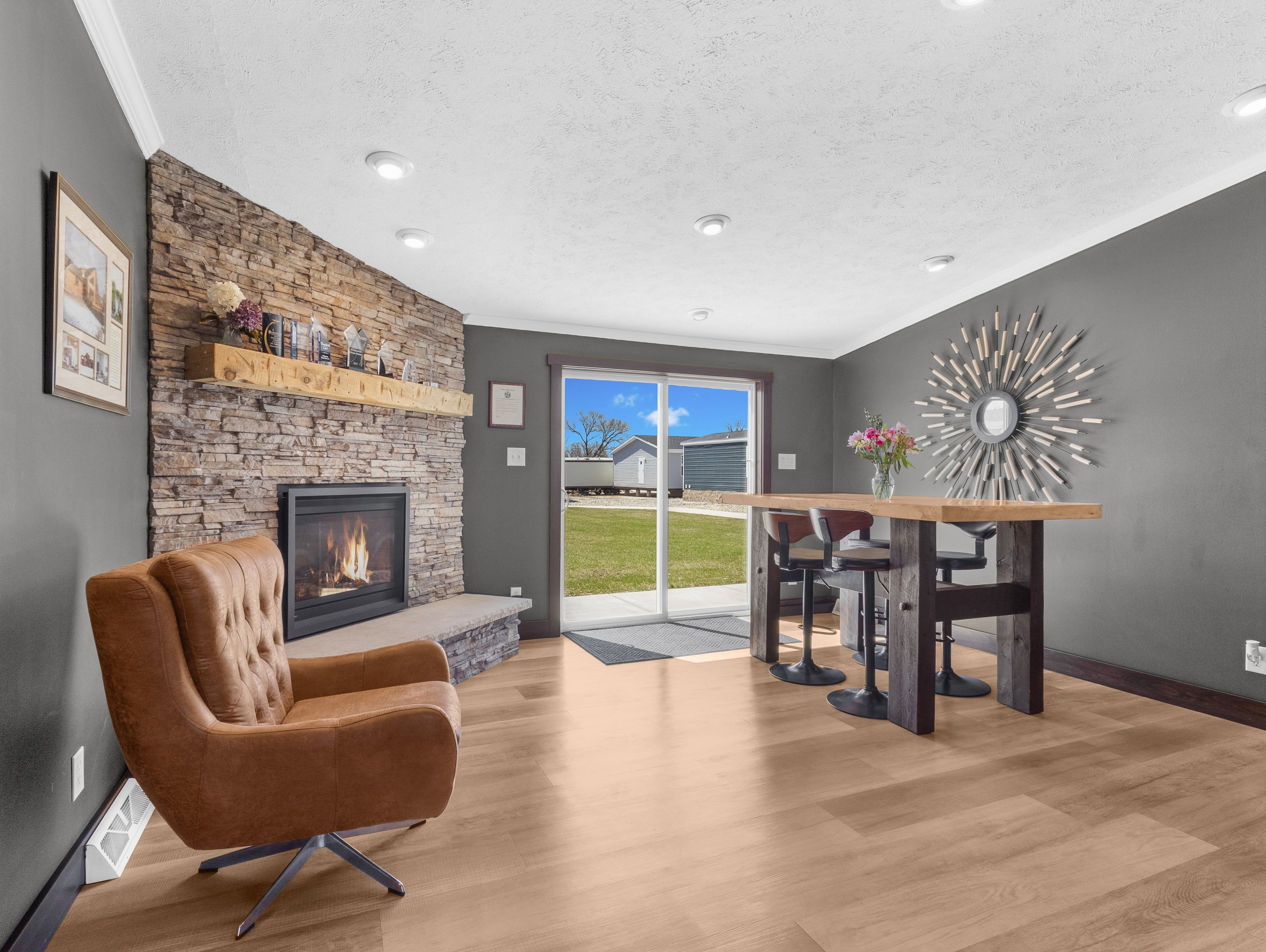 Living room with brown leather armchair, stone fireplace with wooden mantel, gray walls, artwork on wall, sliding glass door showing outdoor view, wall clock with radiating design, flowers on table, ceiling lights.