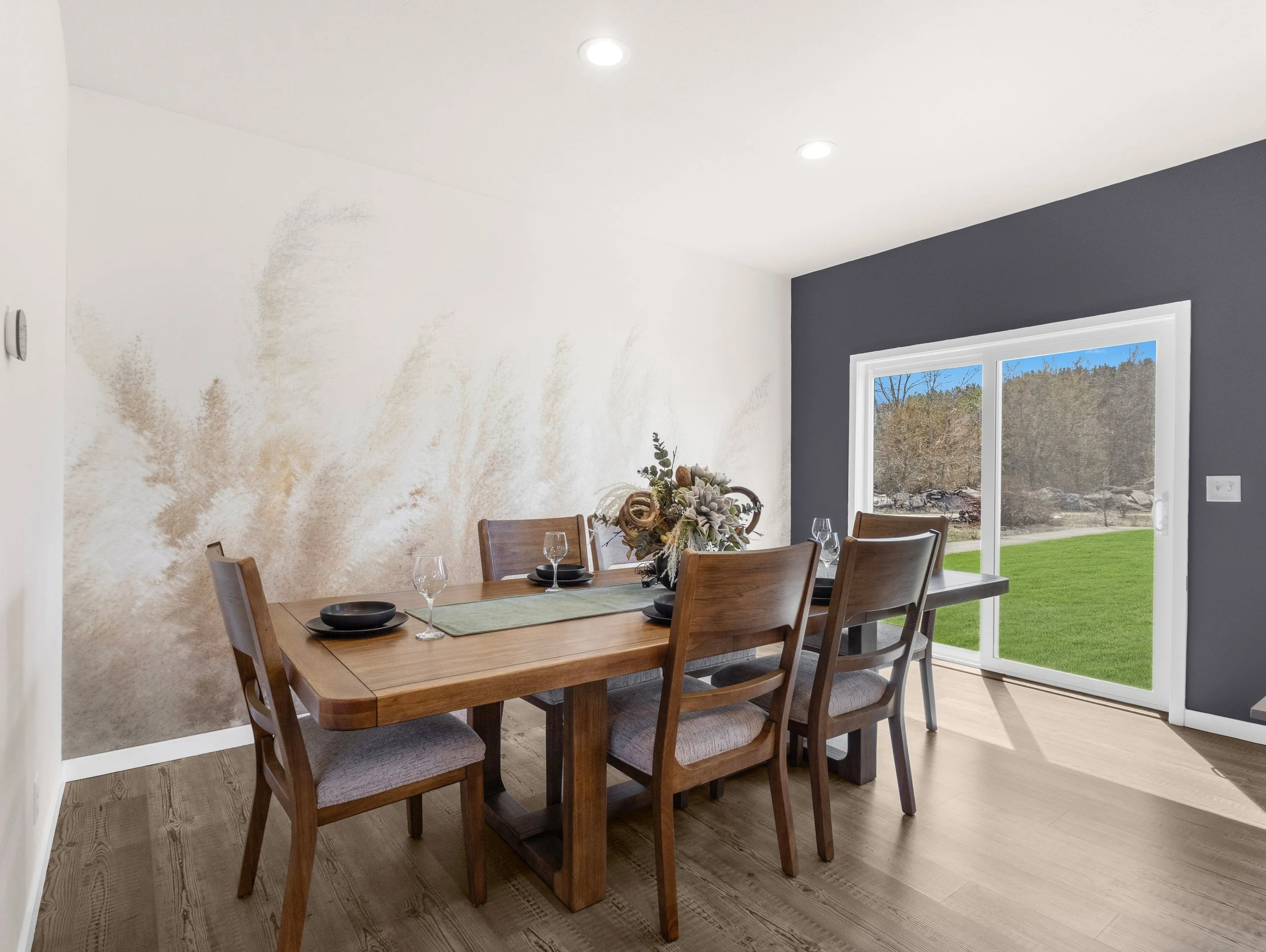 A dining room with a wooden table set with plates, glasses, and a flower arrangement, next to a large window showing a green yard outside. The walls are painted white and dark gray, with recessed ceiling lights.