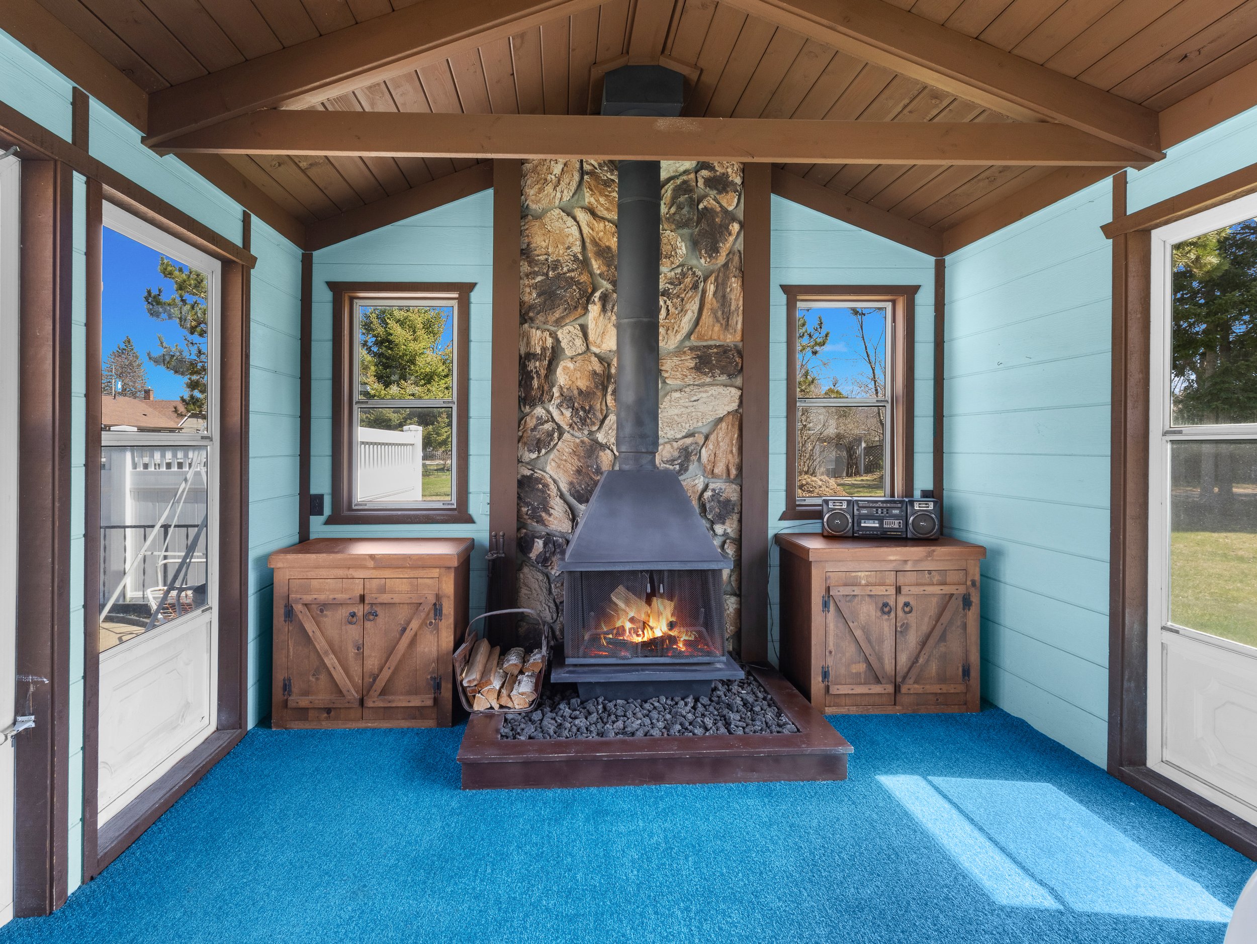 Sunken living room with blue carpet, wooden cabinets, wood-paneled ceiling, a stone fireplace with a wood stove, and windows showing trees outside.