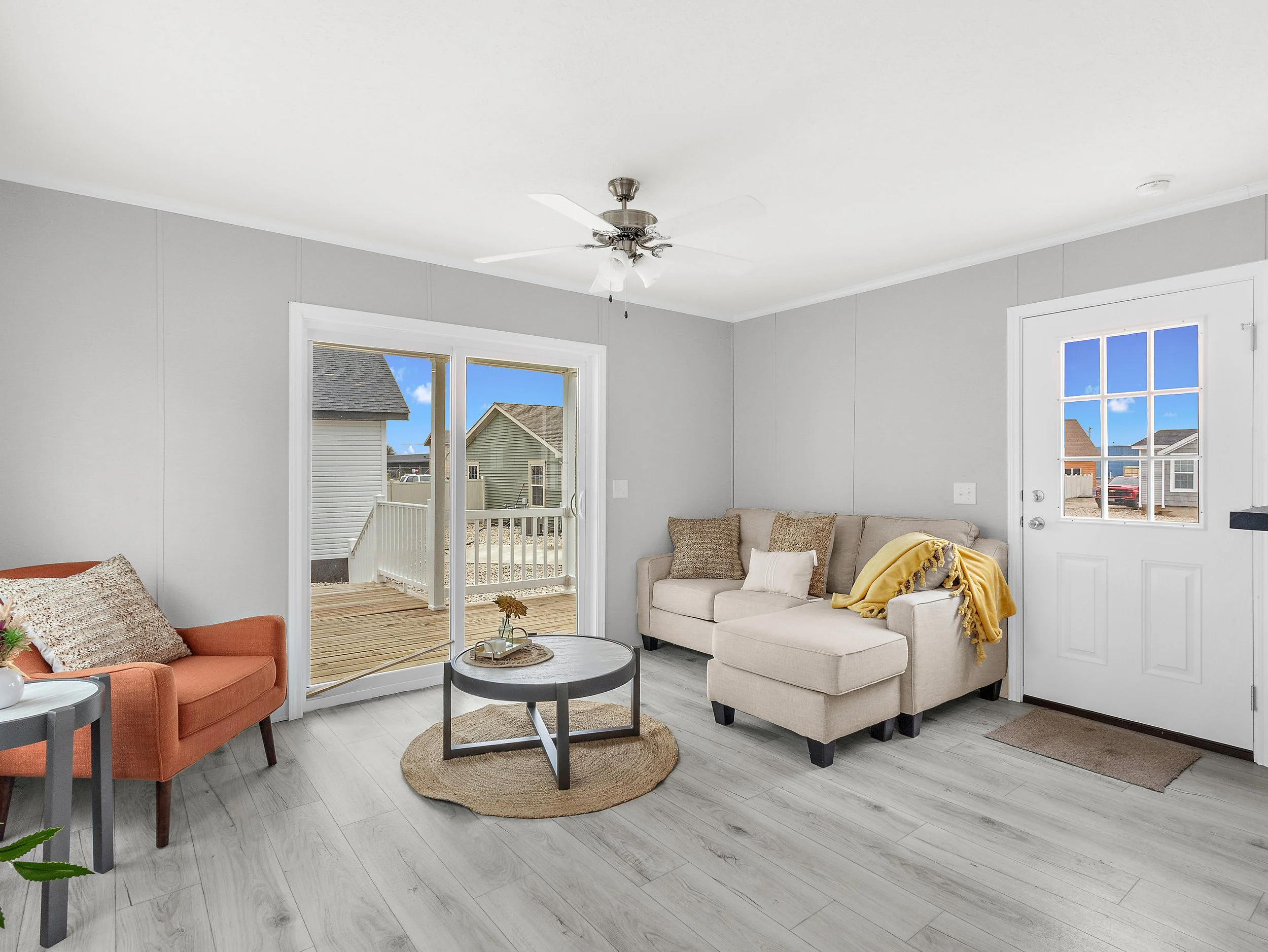Living room with gray walls, light wood flooring, a beige sectional sofa with throw pillows, an orange armchair, a small round coffee table, and sliding glass door leading to a deck with neighboring houses visible outside.