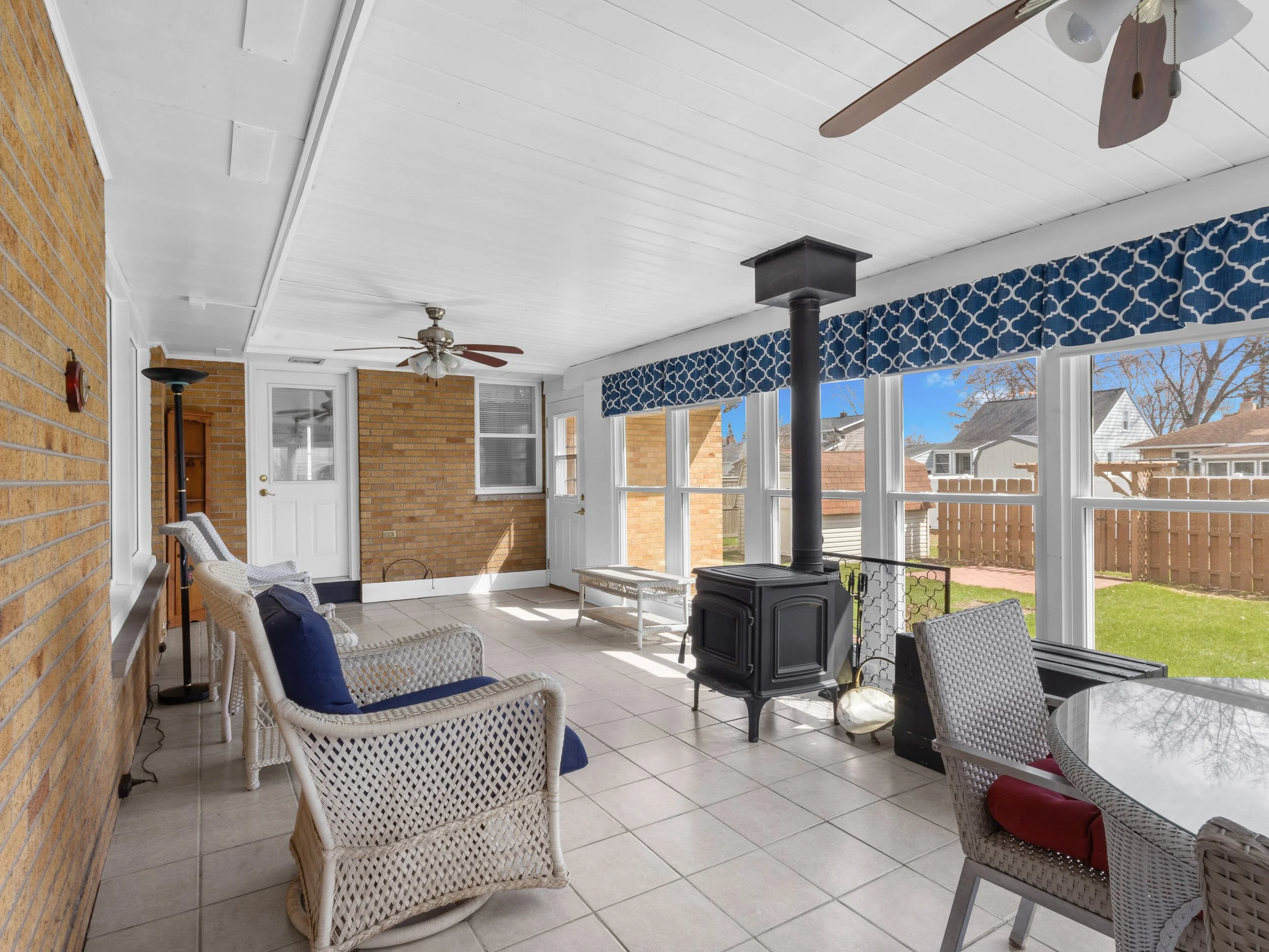 Sunroom with wicker chairs with navy cushions, a black wood stove, ceiling fans, blue patterned curtains, large windows showing a backyard with trees and neighboring houses.
