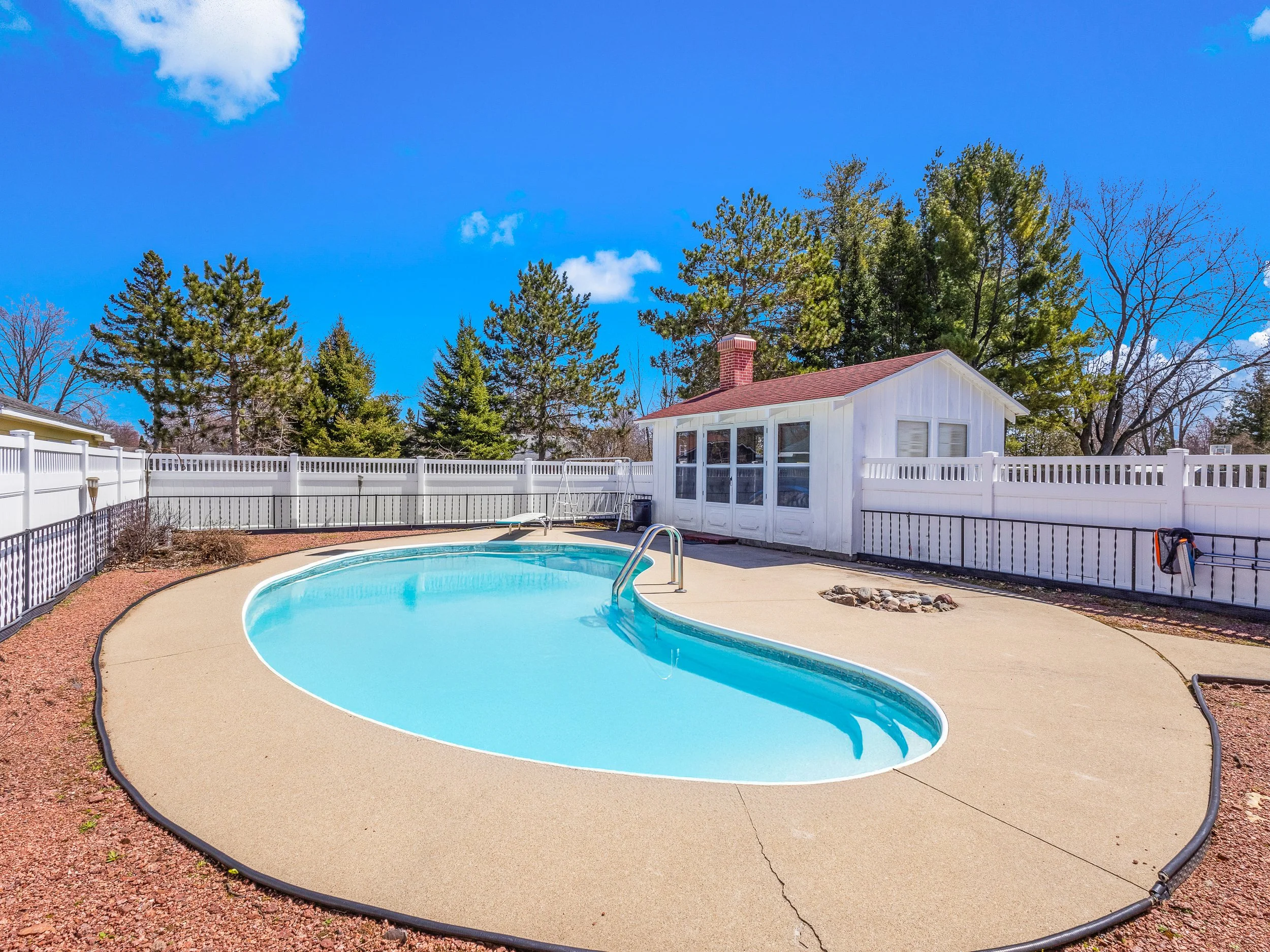 A backyard with a swimming pool, white fence, and a small white shed, surrounded by trees and under a partly cloudy blue sky.