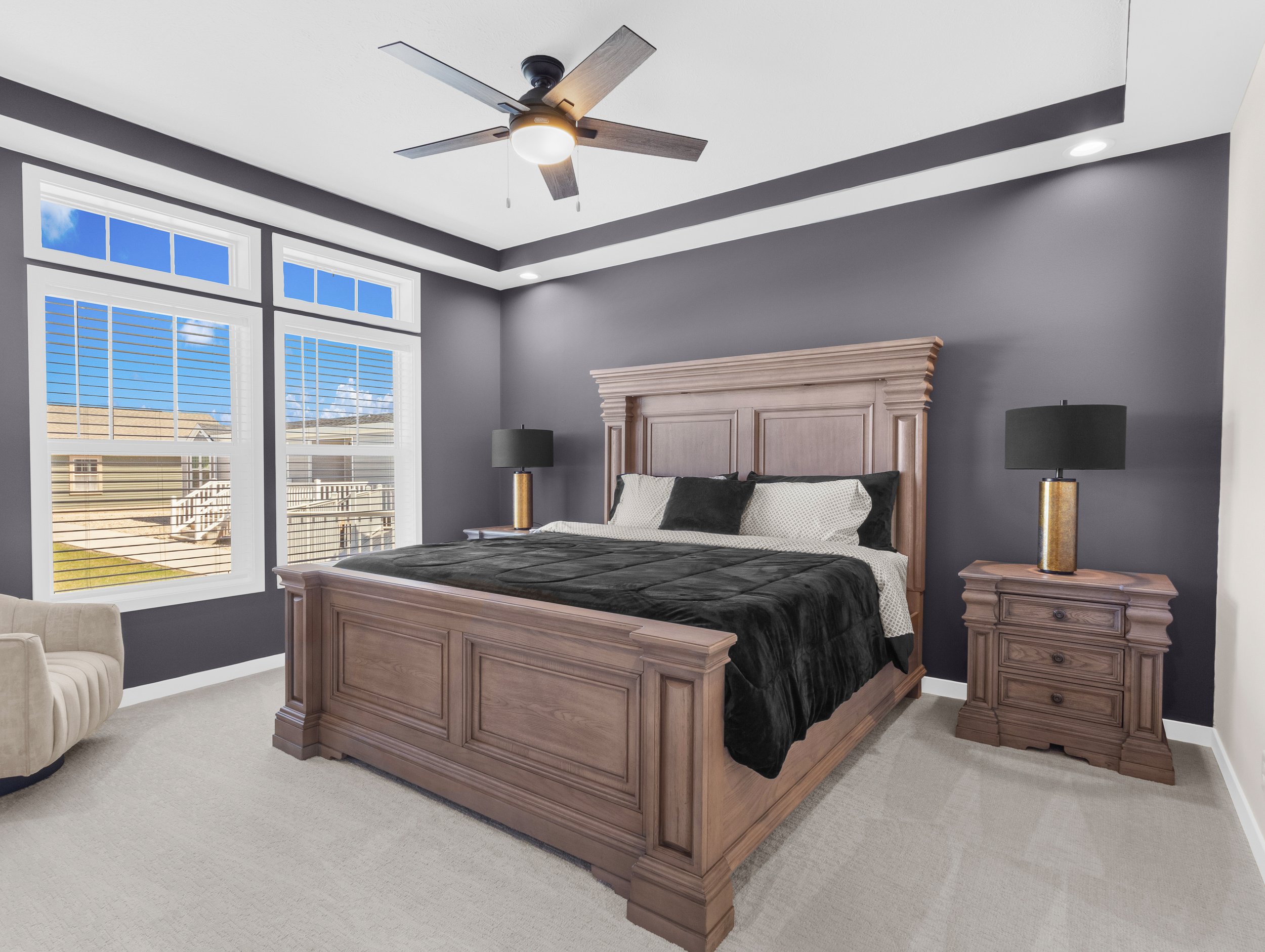 A bedroom with gray walls, a large window with blinds showing houses and blue sky outside, a wooden bed frame with matching nightstands, black and white bedding, two black lamps on gold stands, a ceiling fan with five wooden blades and a light, and a