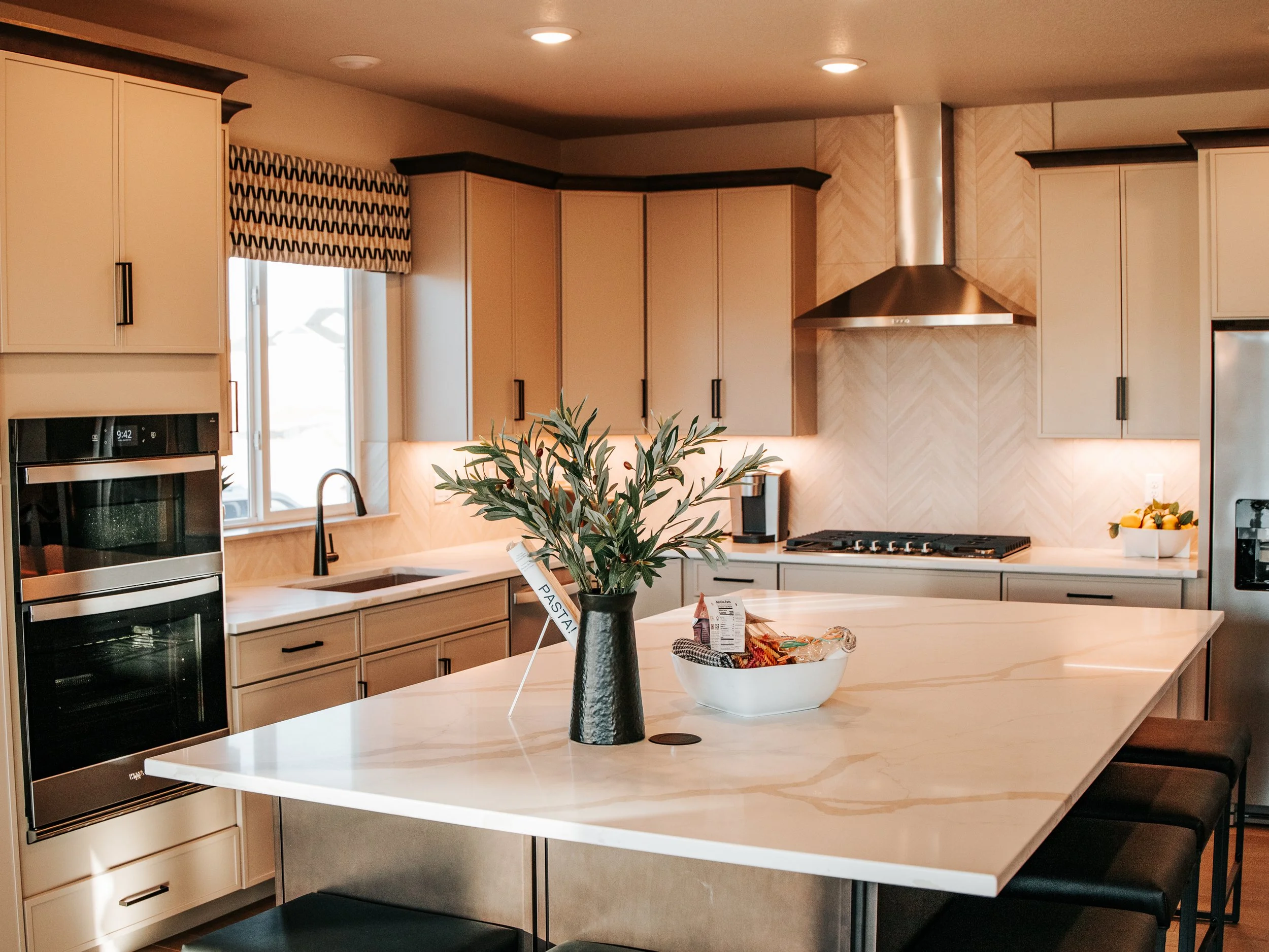 Modern kitchen with white countertops, beige cabinets, black hardware, stainless steel appliances, a plant on the island, and a window with a patterned valance.
